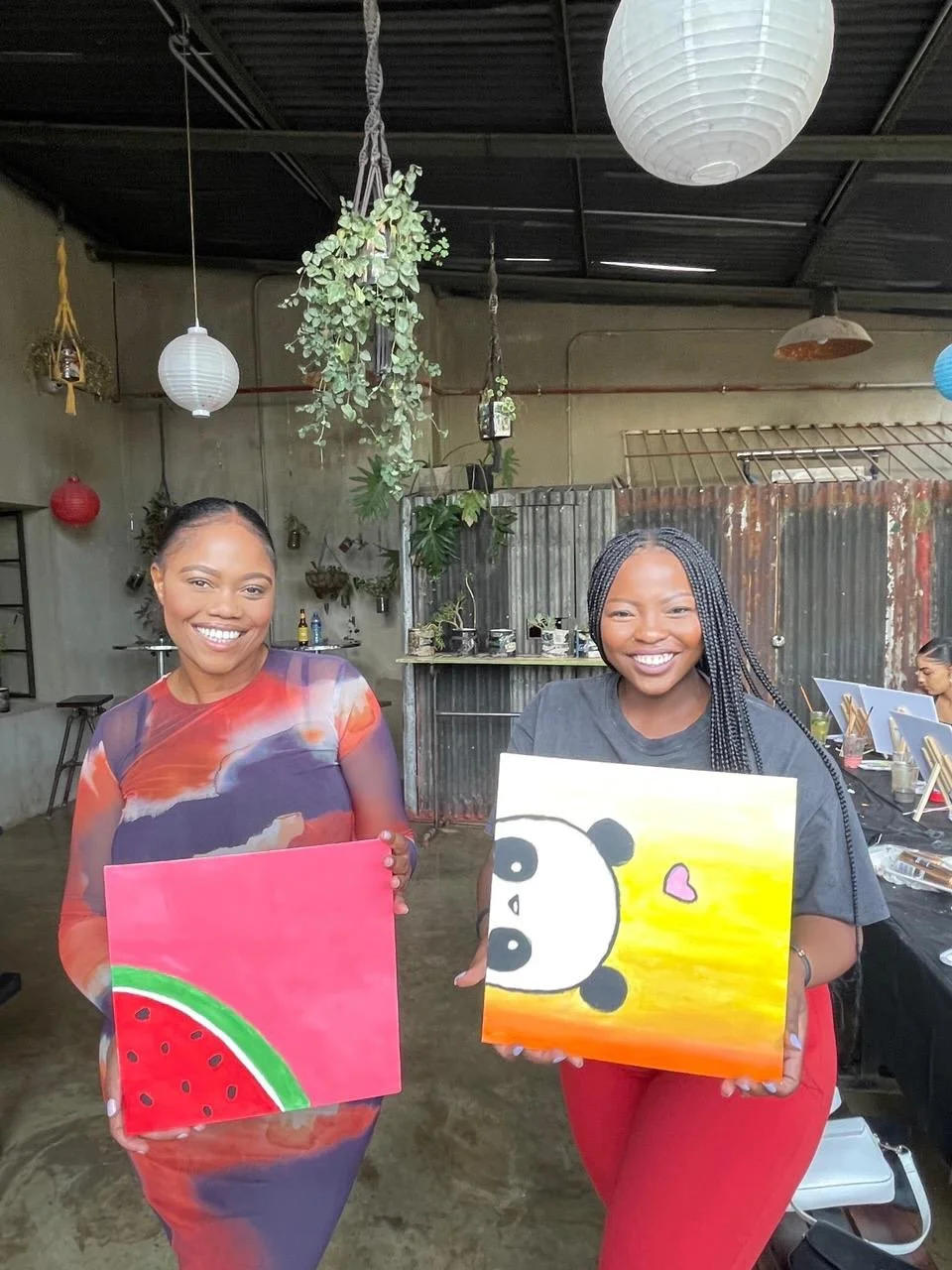 Two women smiling and showing their colorful paintings, one with a watermelon and the other with a panda and a pink heart, inside an art studio.
