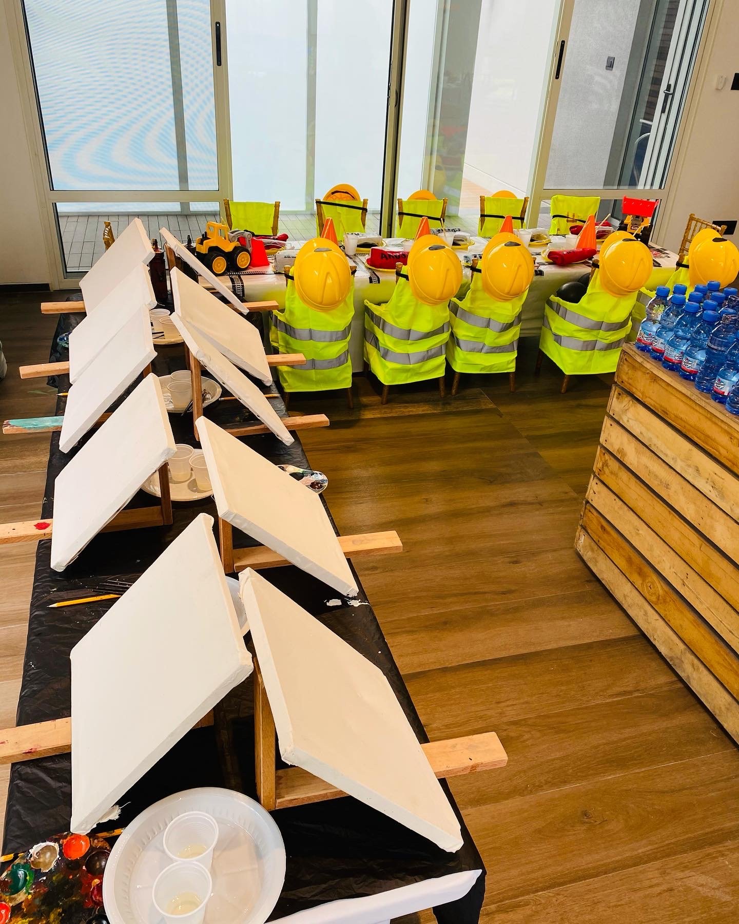 A table set for children with toy construction helmets, toy trucks, and toy tools, likely for a themed birthday party or activity. There are empty canvases and art supplies nearby.