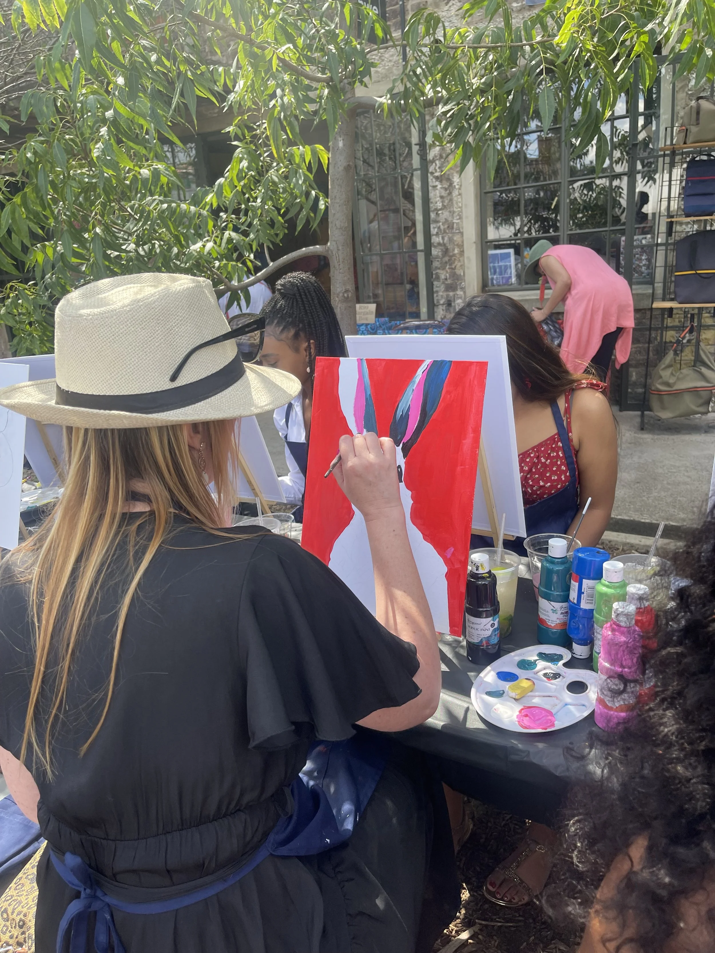 People painting floral artwork at an outdoor event with art supplies and a large tree in the background.