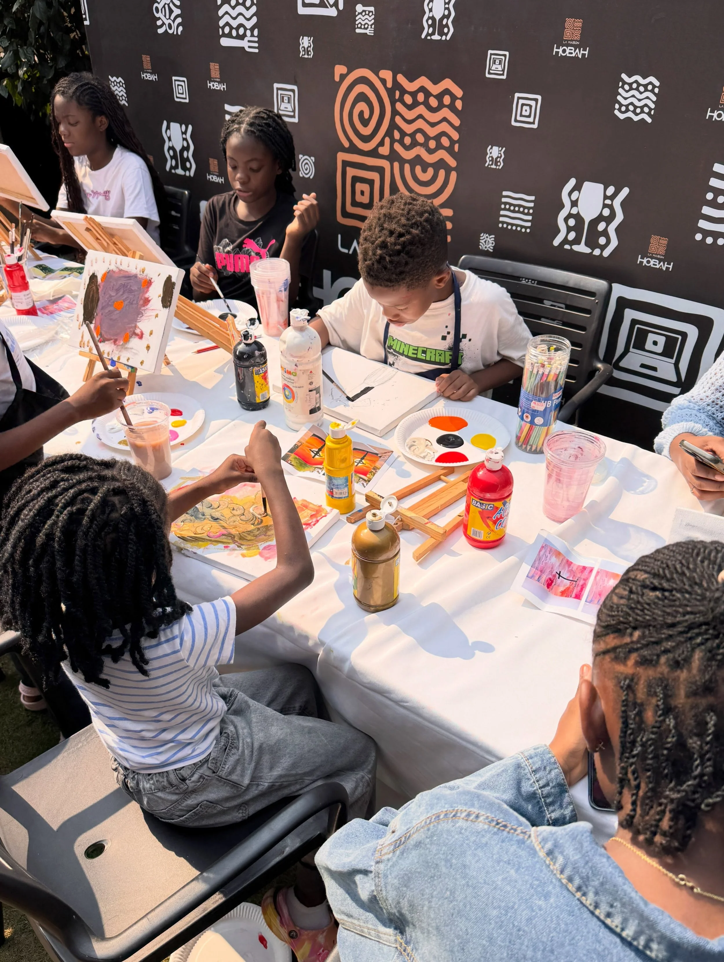 Children seated at a table outdoors, painting on canvases with various art supplies such as paints, brushes, and paper. A black backdrop with white and orange geometric designs and the word 'HOBAN' is visible behind them.