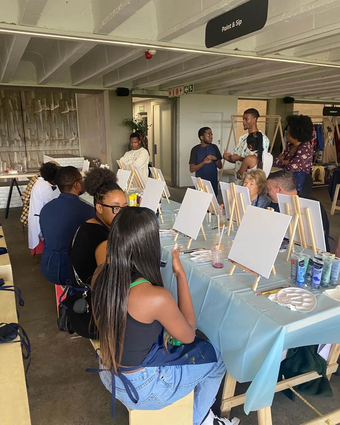A group of people participating in a painting class, sitting around a table with blank canvases and paint supplies, under a sign that says 'Paint & Sip'.