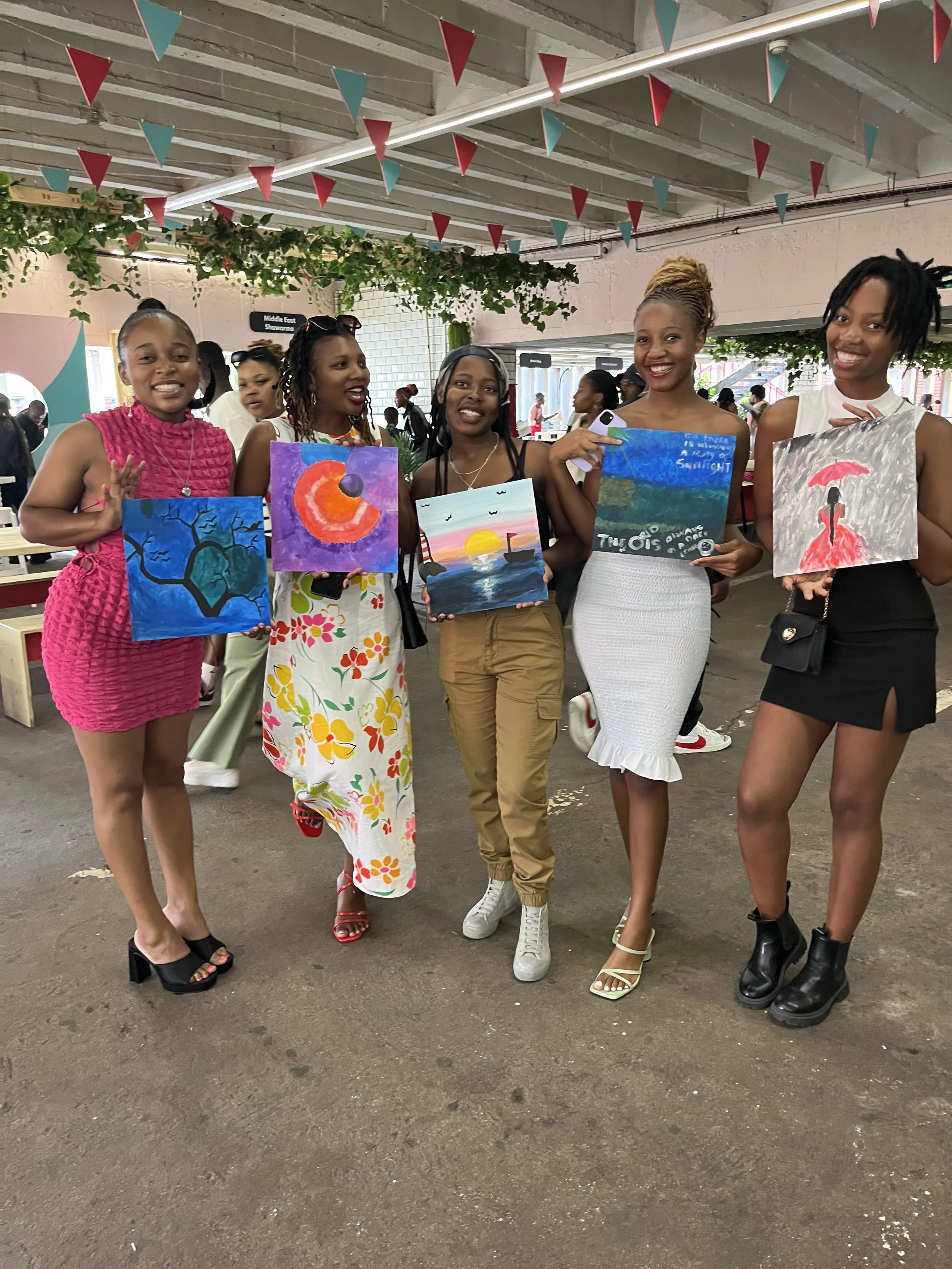 Five women standing together and smiling, holding colorful paintings, at an indoor event with decorations and people in the background.