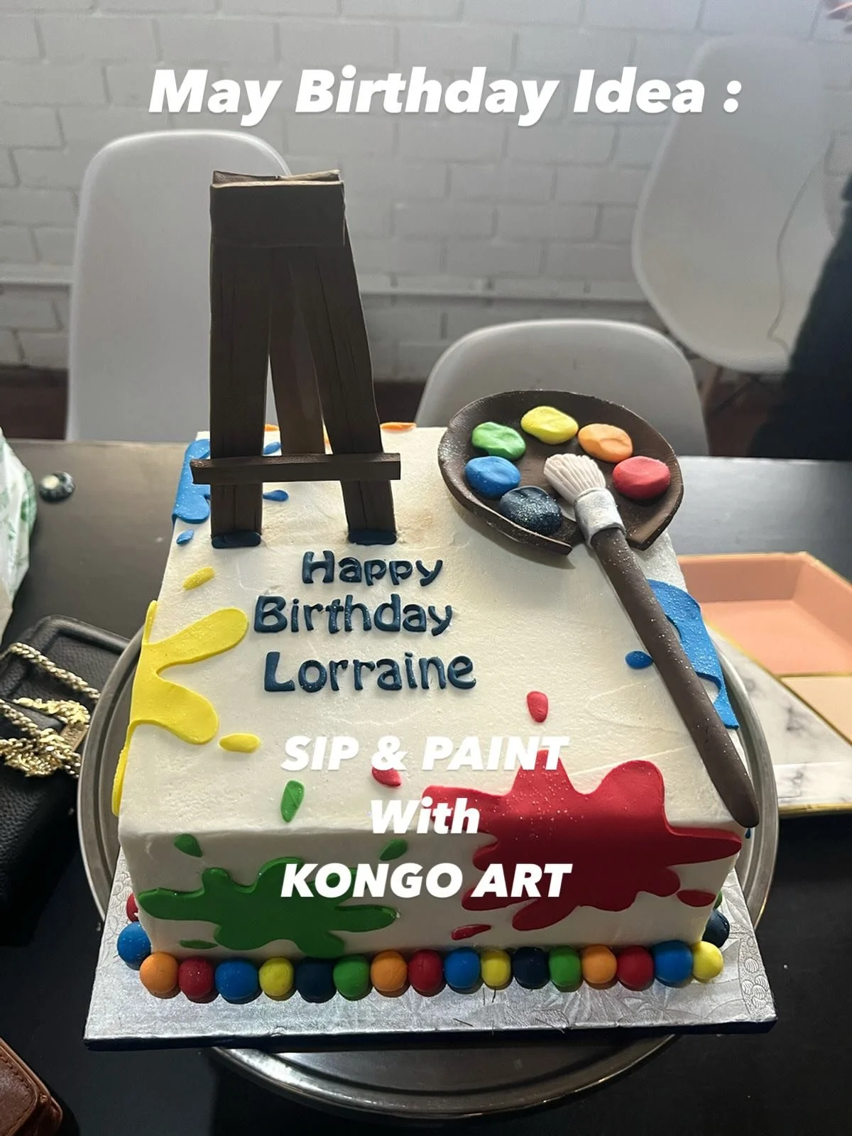 A birthday cake with white icing and colorful splatters, decorated with a fondant paint palette, paintbrush, and easel topper. The cake has blue and yellow text that says "Happy Birthday Lorraine" and additional text that reads "SIP & PAINT With KONGO ART."