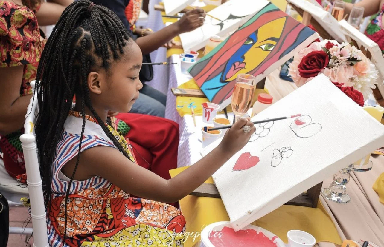A young girl with braided hair painting on a canvas with a heart and flower designs. She is seated at a decorated table with flowers, drinks, and other artworks, and is focused on her painting.