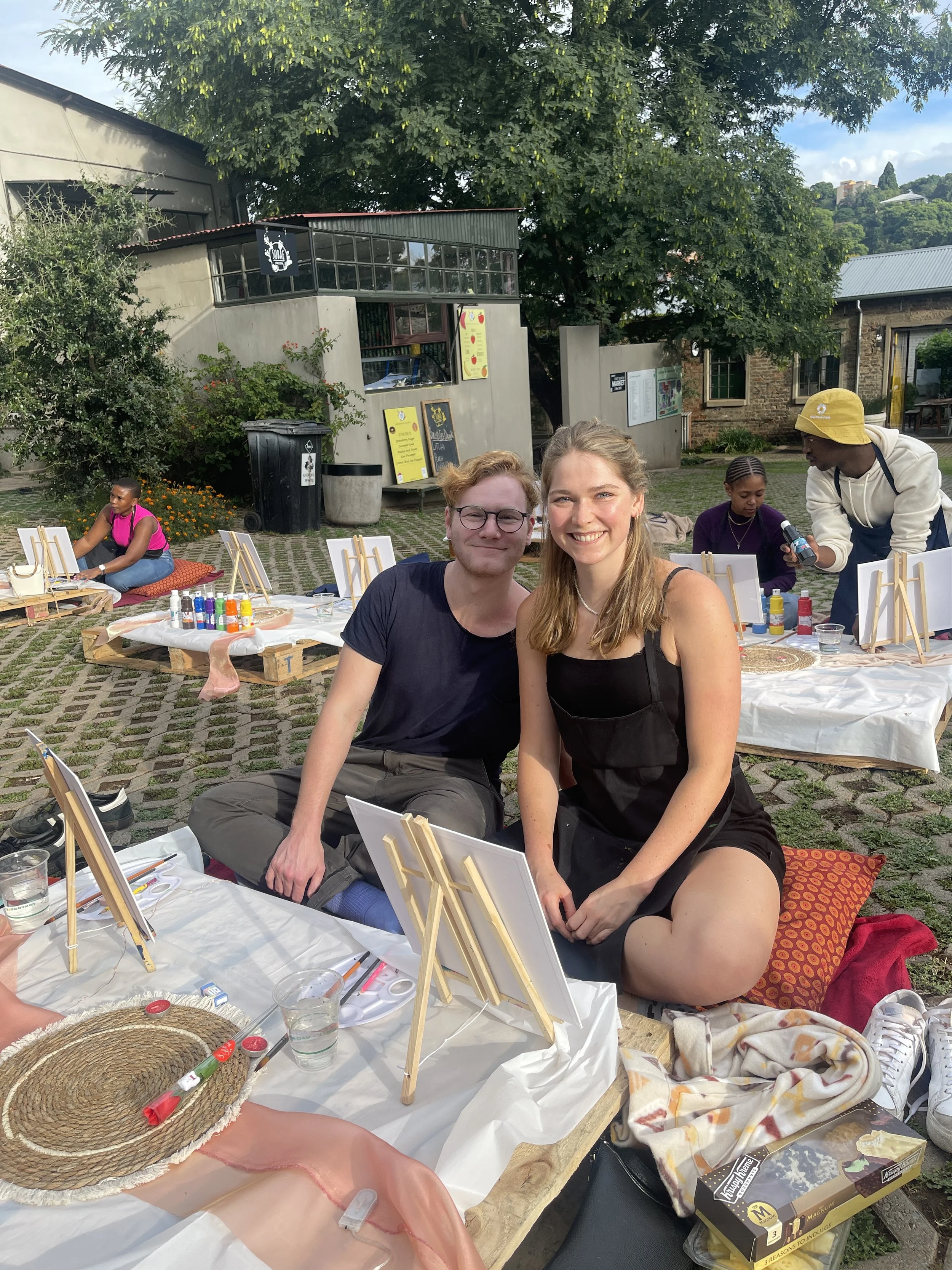 An outdoor art activity session with two young adults sitting together, smiling at camera. There are painting supplies on the table, including small canvases, paints, brushes, and easels. In the background, other people are engaged in painting activi