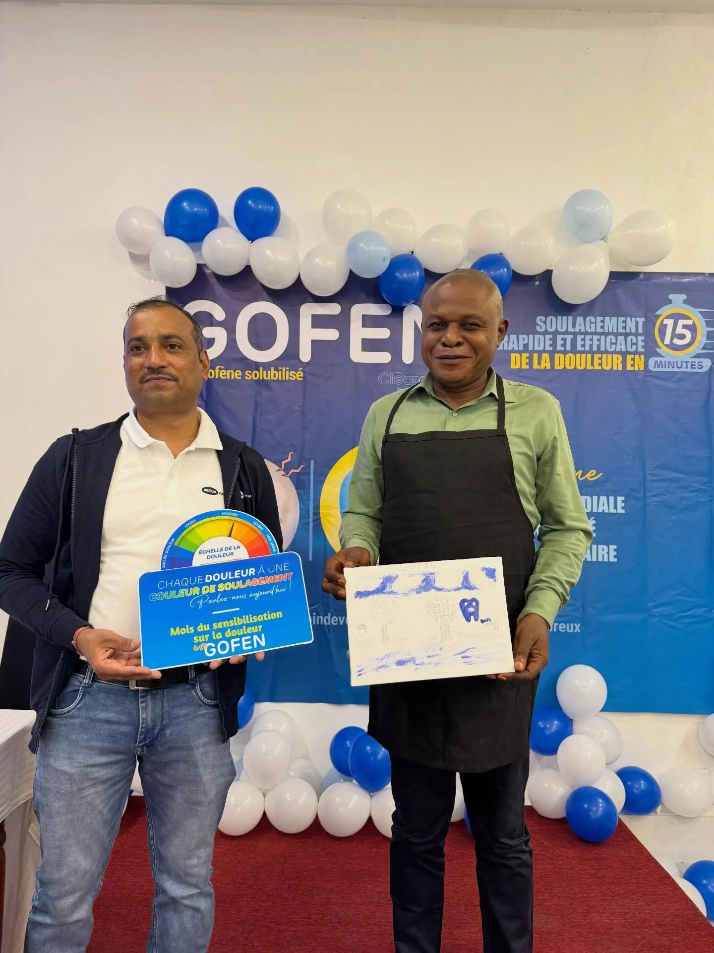 Two men standing in front of a blue and white balloon backdrop with the words 'Gofen' and additional French text. One man is holding a blue sign related to pain relief and the other is holding a white board with blue markings. The scene appears to be