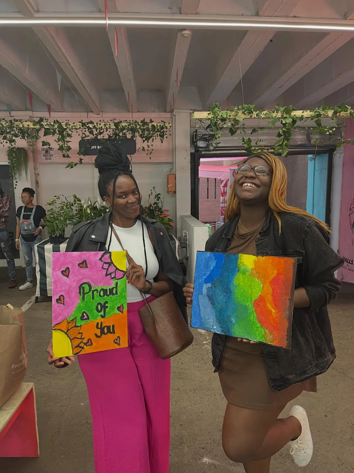 Two women smiling and holding colorful artwork at an indoor event. The woman on the left holds a sign that reads "Proud of You" with hearts and sun drawings. The woman on the right holds a rainbow-colored painting, kneeling on one knee, wearing glass