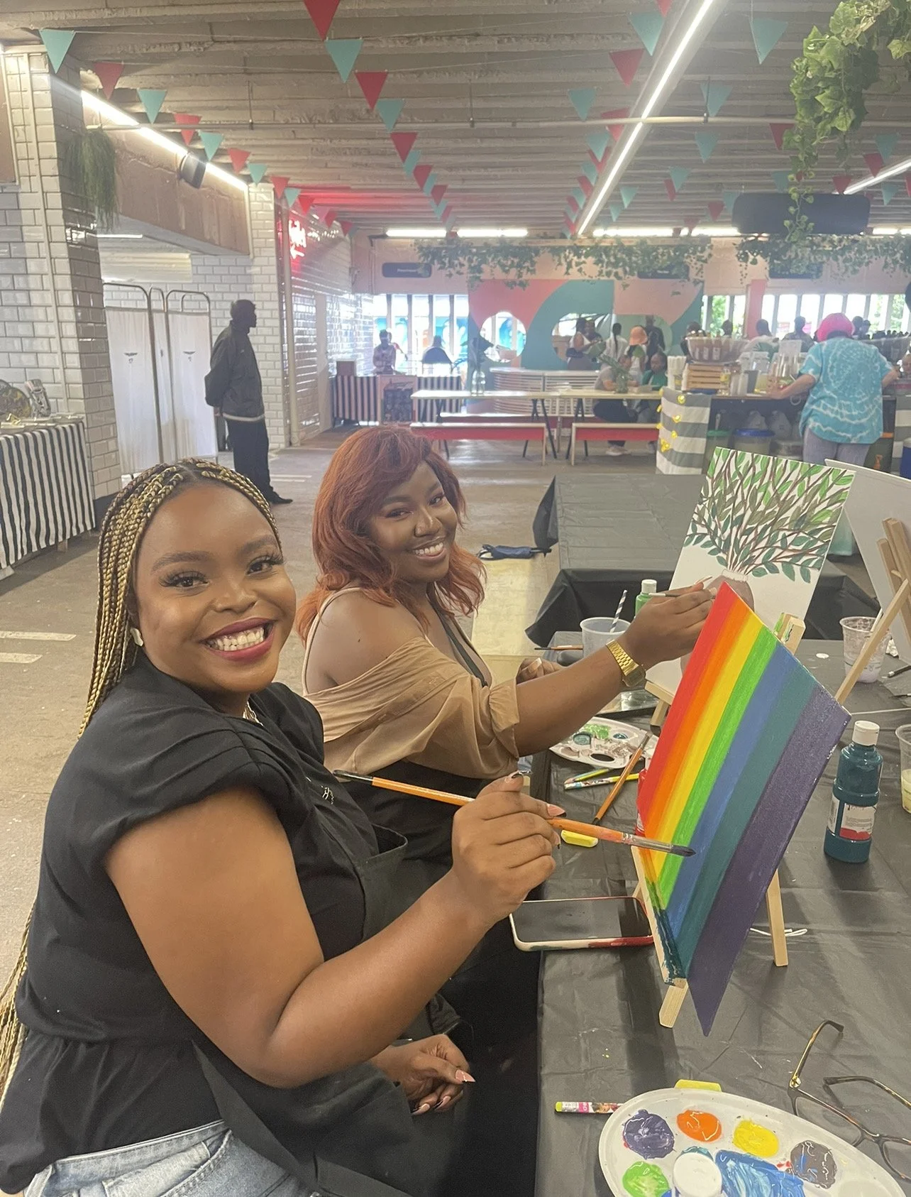 Two women smiling and painting rainbow-colored canvases at an indoor art event, with other people and decorations visible in the background.