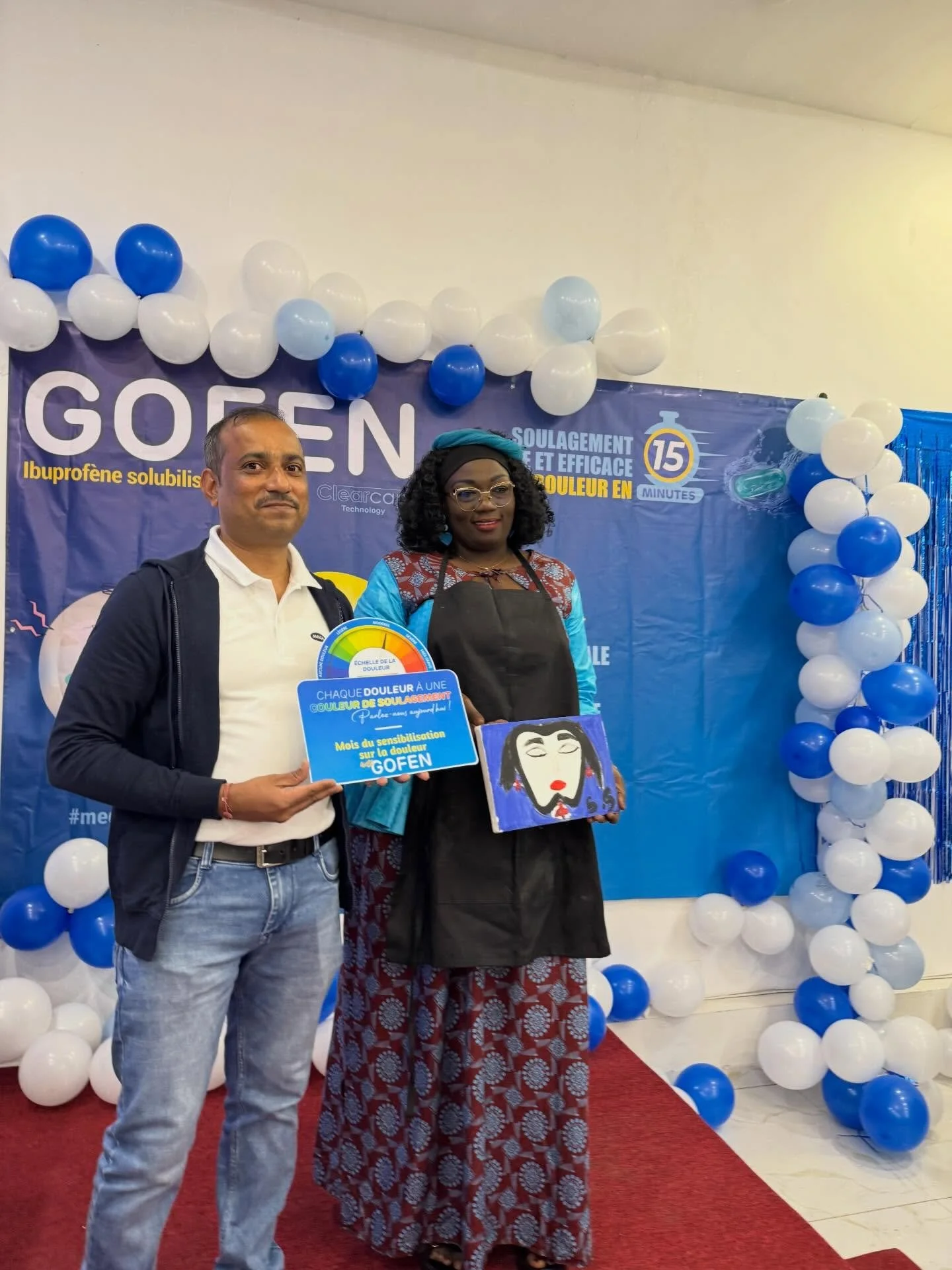 A man and woman standing on a red carpet, holding signs, with a backdrop decorated with white and blue balloons and a purple banner promoting pain relief with Gofen.