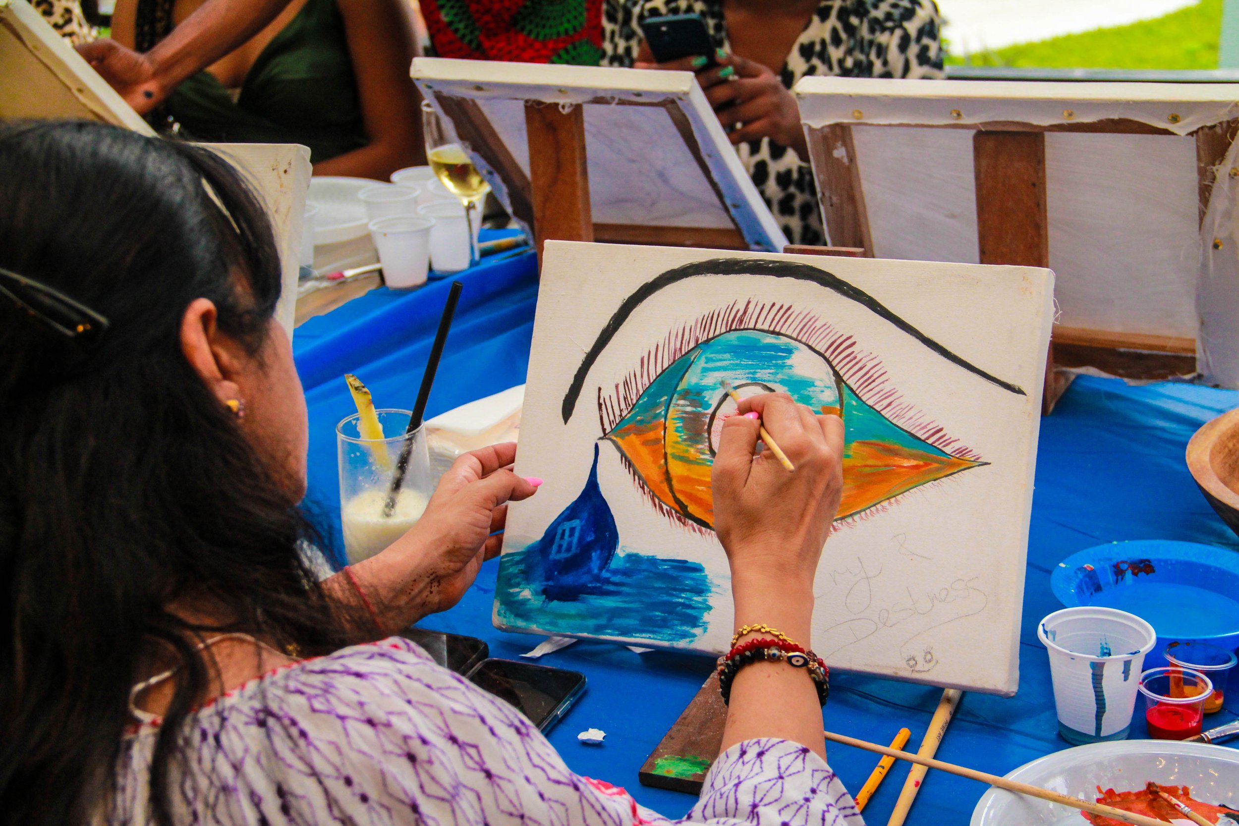 A person painting a colorful canvas of an eye with a landscape reflection inside it at an outdoor art event. Other people and art supplies are visible in the background.