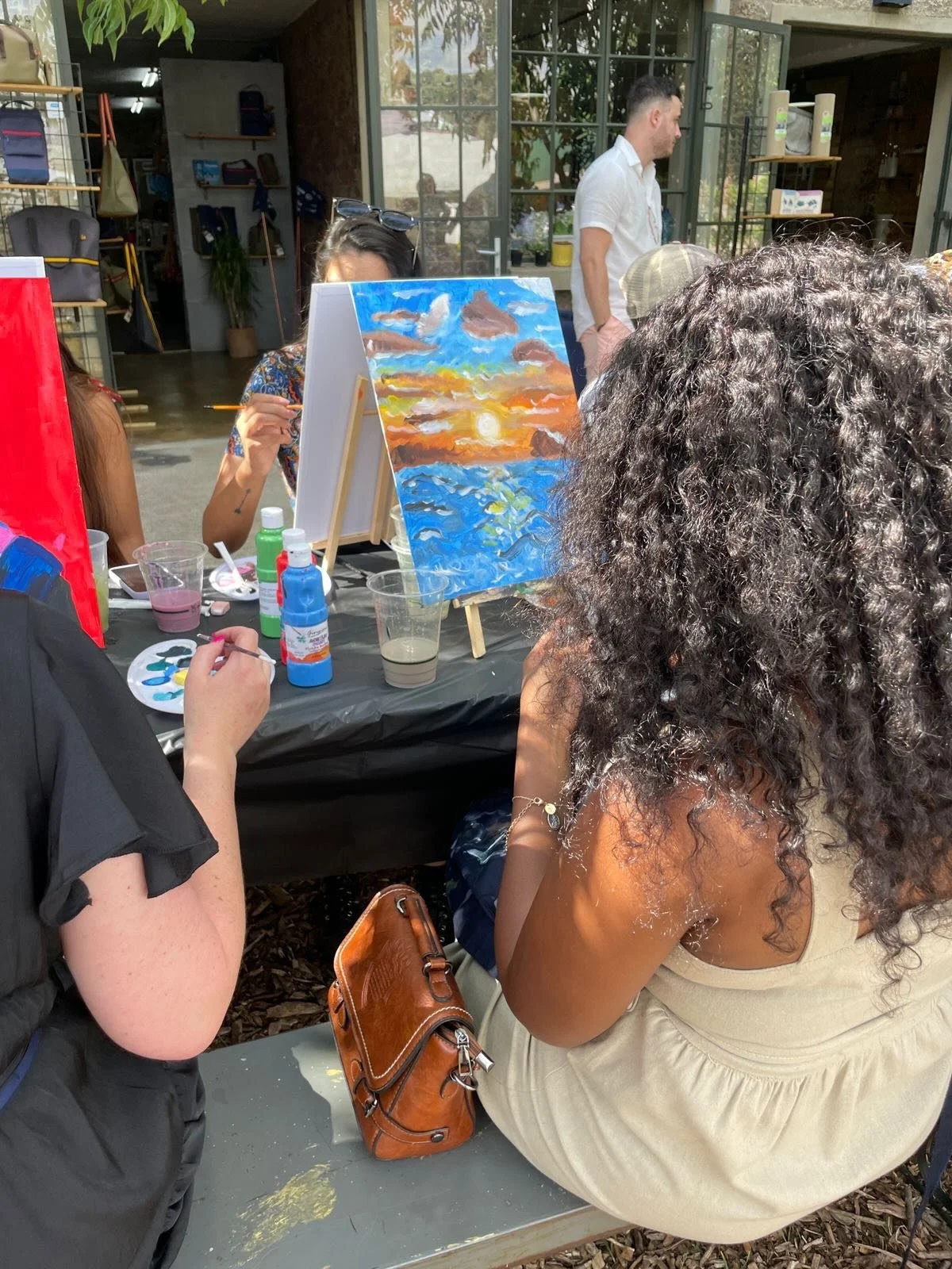 People painting outdoor scene on canvases at a group paint session. One woman with curly hair is seated with her back to the camera, another woman is painting with sunglasses on her head, and a man is standing indoors near shelves with bags. Painting