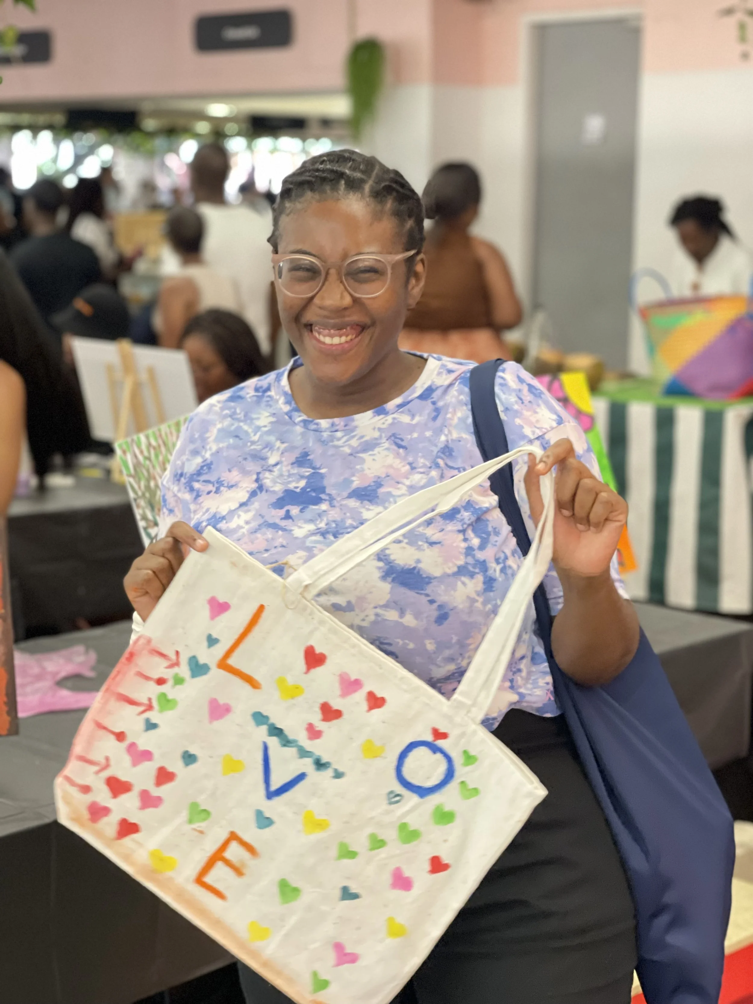 A woman smiling and holding a colorful, decorated paper bag at an indoor event with other people and vendor tables in the background.