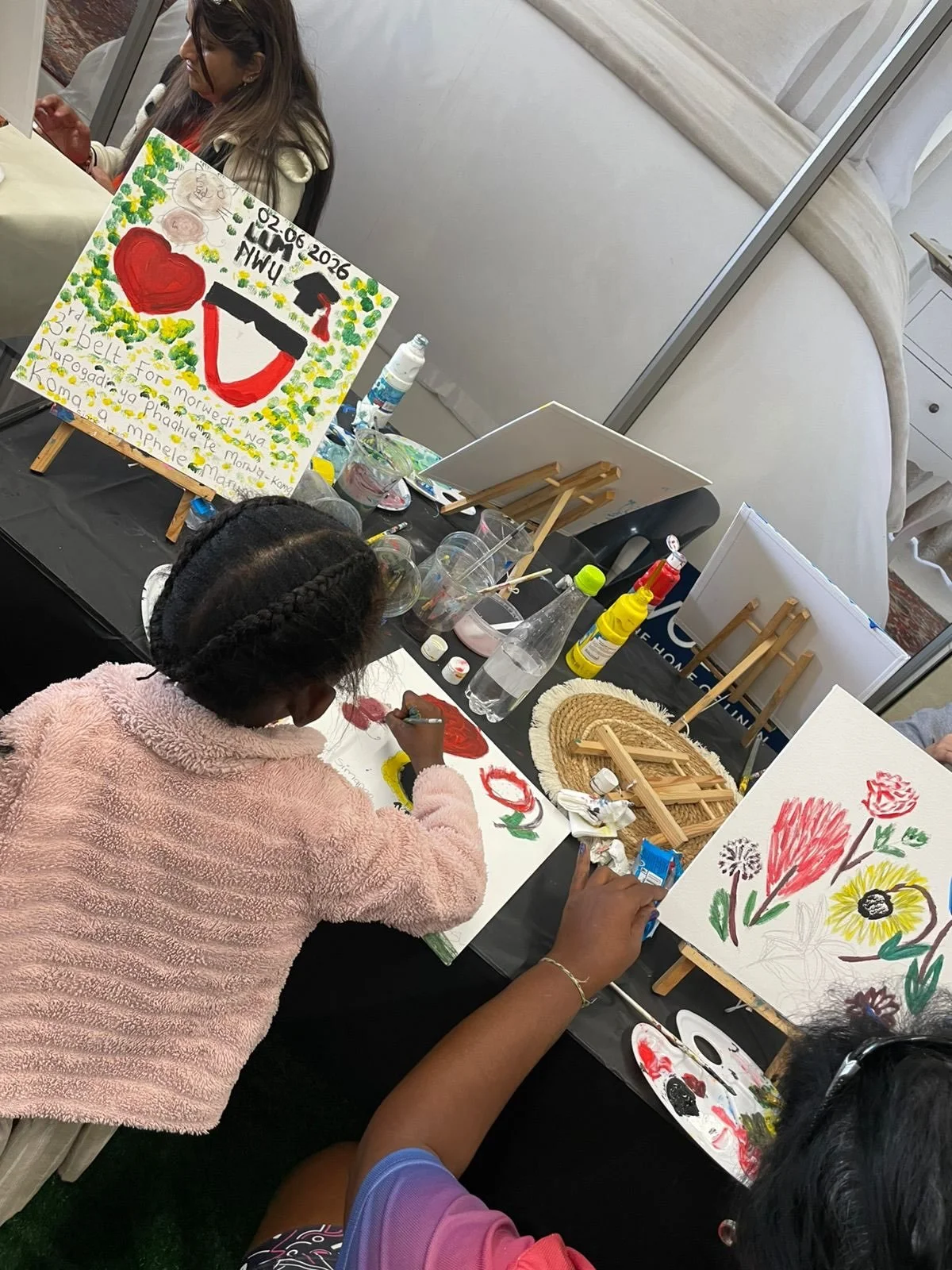 Children and adults participating in a painting activity at a table covered with art supplies, including paint, brushes, and water bottles. There are completed and in-progress paintings of flowers and a message on display.