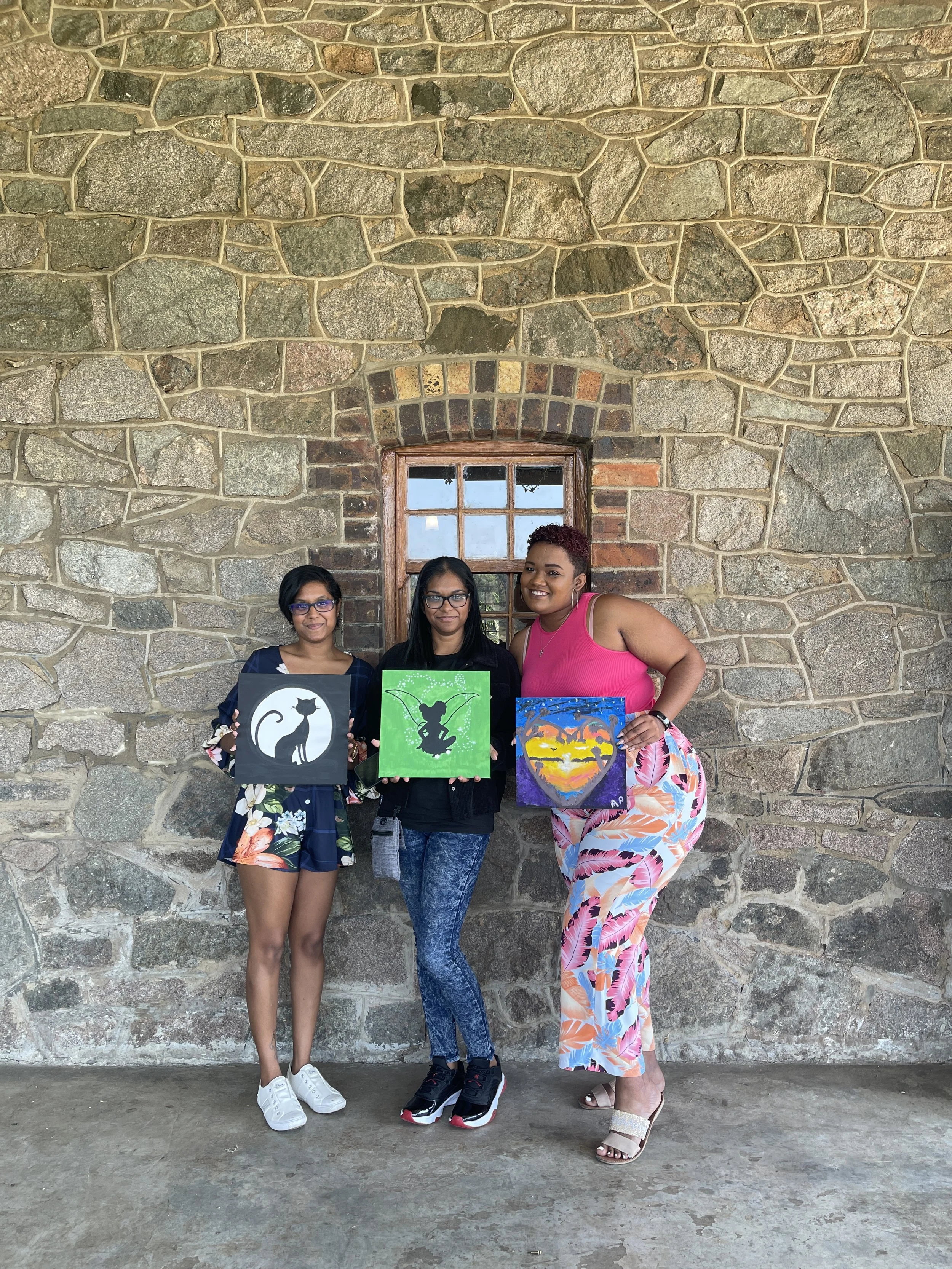 Three women standing in front of a stone wall with a small window, holding colorful paintings of cats, bats, and a sunset.