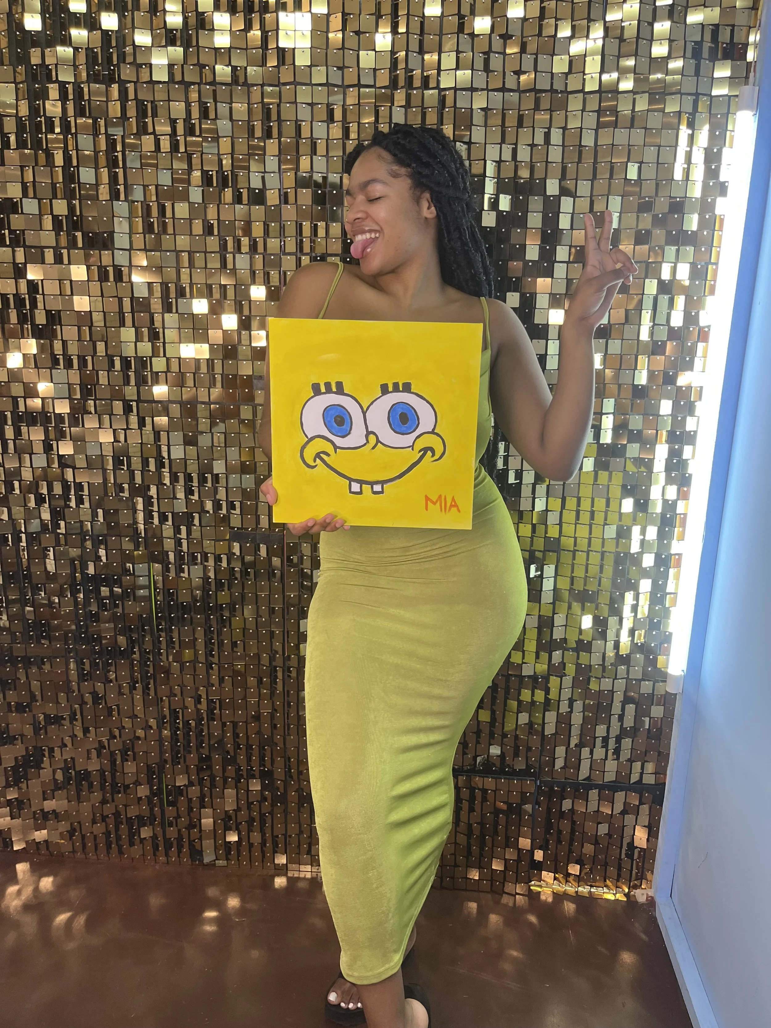 Young woman with long dreadlocks wearing a green dress holding a yellow painting of SpongeBob SquarePants and posing with her eyes closed and tongue out, standing in front of a shiny, metallic mosaic wall.