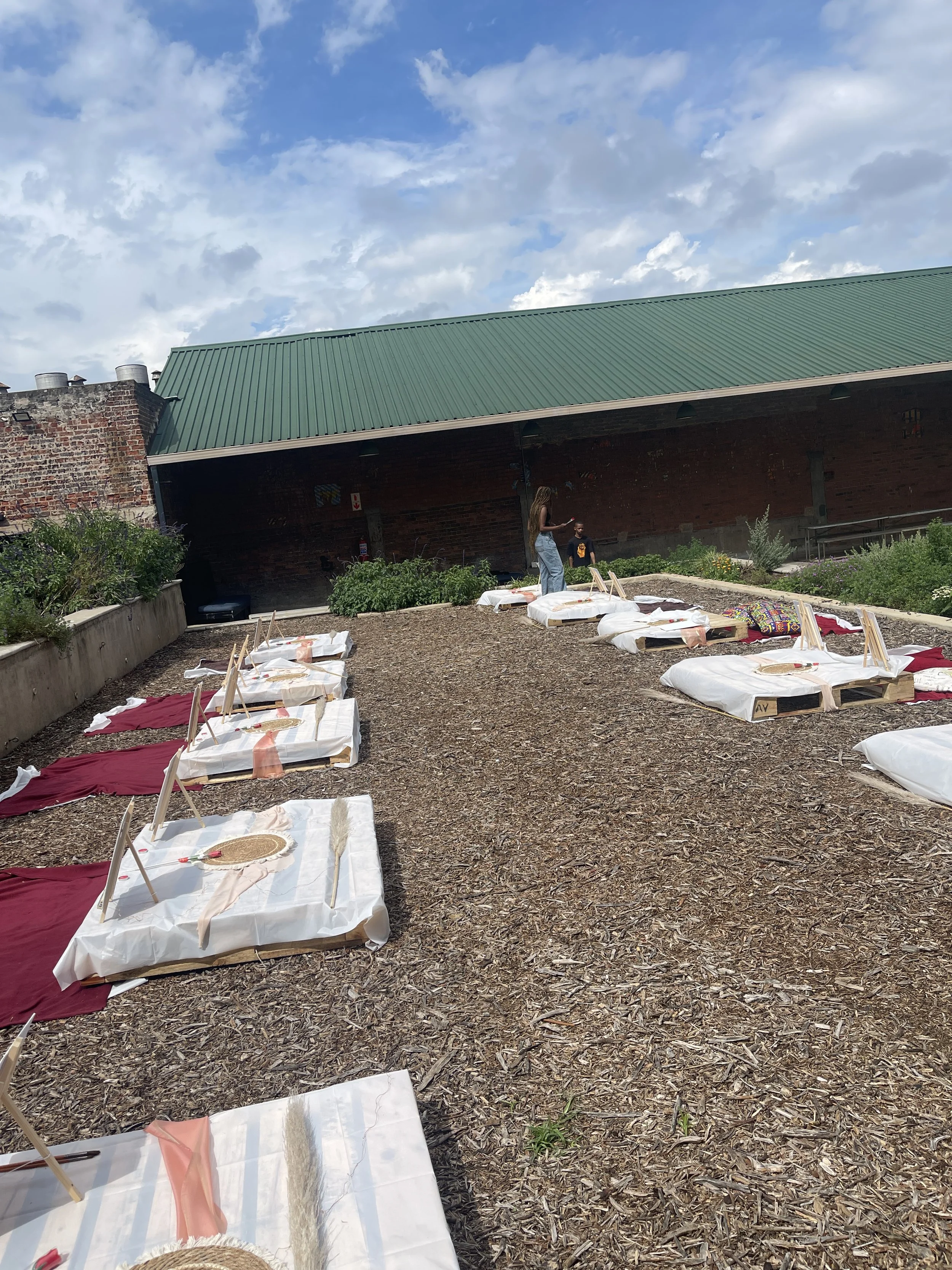 Outdoor space with several white and red cloth-covered platform beds, some with wooden frames, set on a ground covered with wood chips. Two people are standing near a brick wall with a green metal roof. There are plants and bushes along one side, und