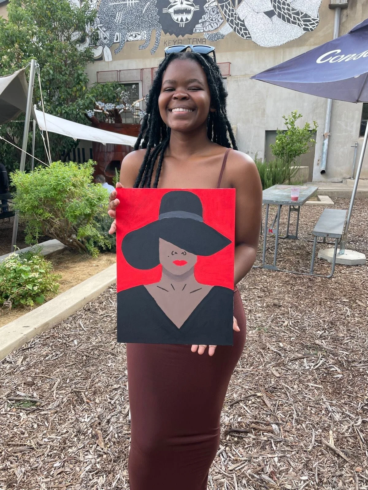 A woman with braided hair and sunglasses on her head smiling and holding a painting of a woman wearing a wide-brimmed hat, red lipstick, with a red background behind her, outdoors in a garden area with trees, bushes, and a mural on a nearby wall.