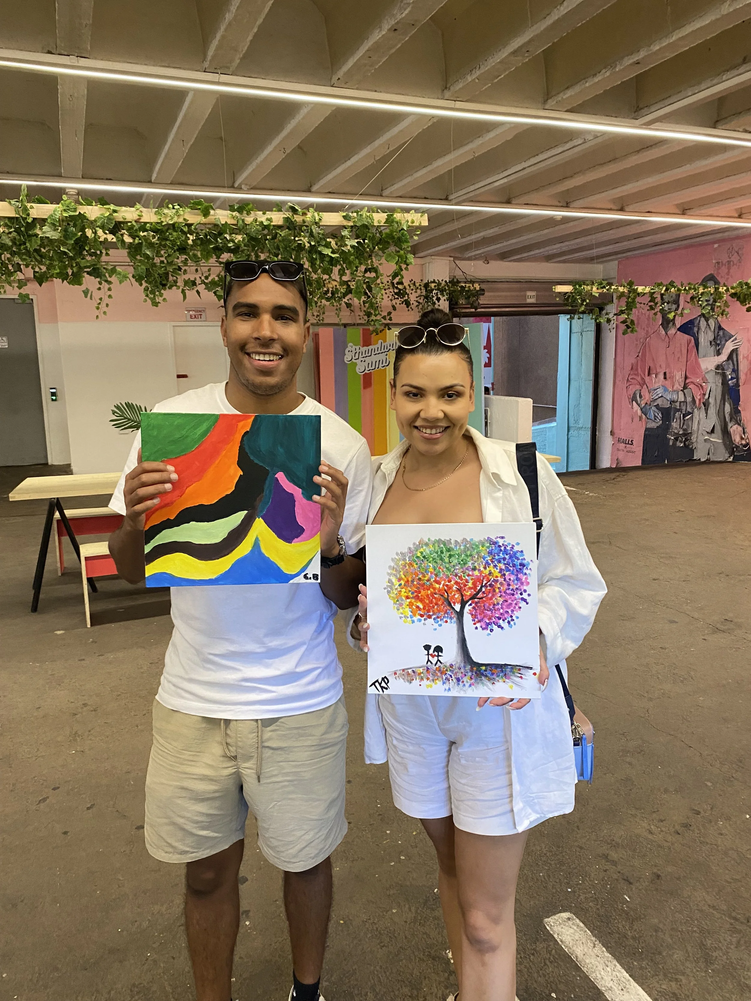 A man and woman standing together in an indoor space, each holding colorful artwork. The man is wearing a white t-shirt, beige shorts, and sunglasses on his head. The woman is wearing a white button-up shirt and white shorts, with sunglasses on her h
