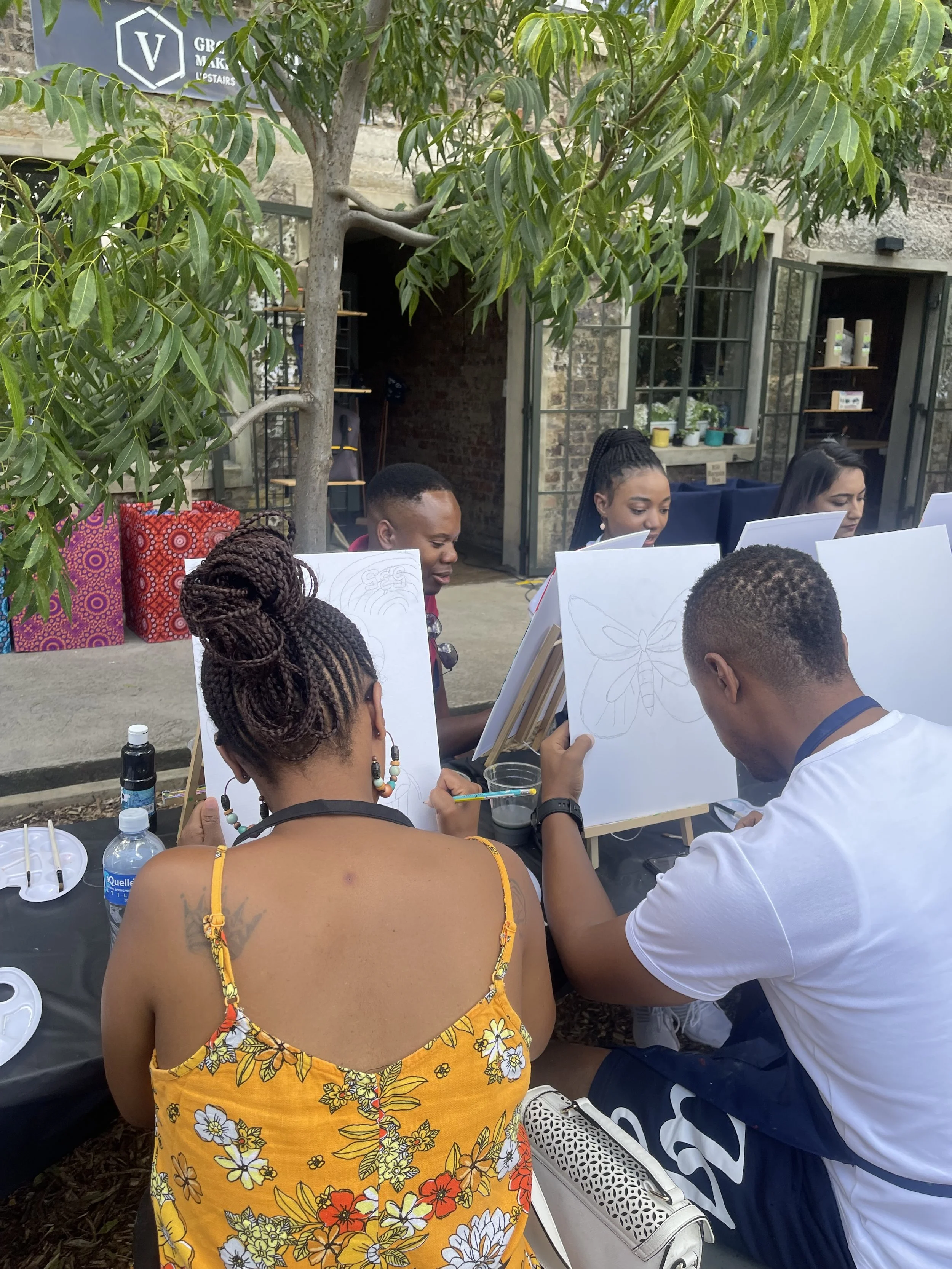 A group of people sitting outdoors around a table, engaged in a drawing activity with floral sketches on paper. A woman in a yellow floral dress is seen from behind, and several others are visible, including a woman with braided hair and a man in a w