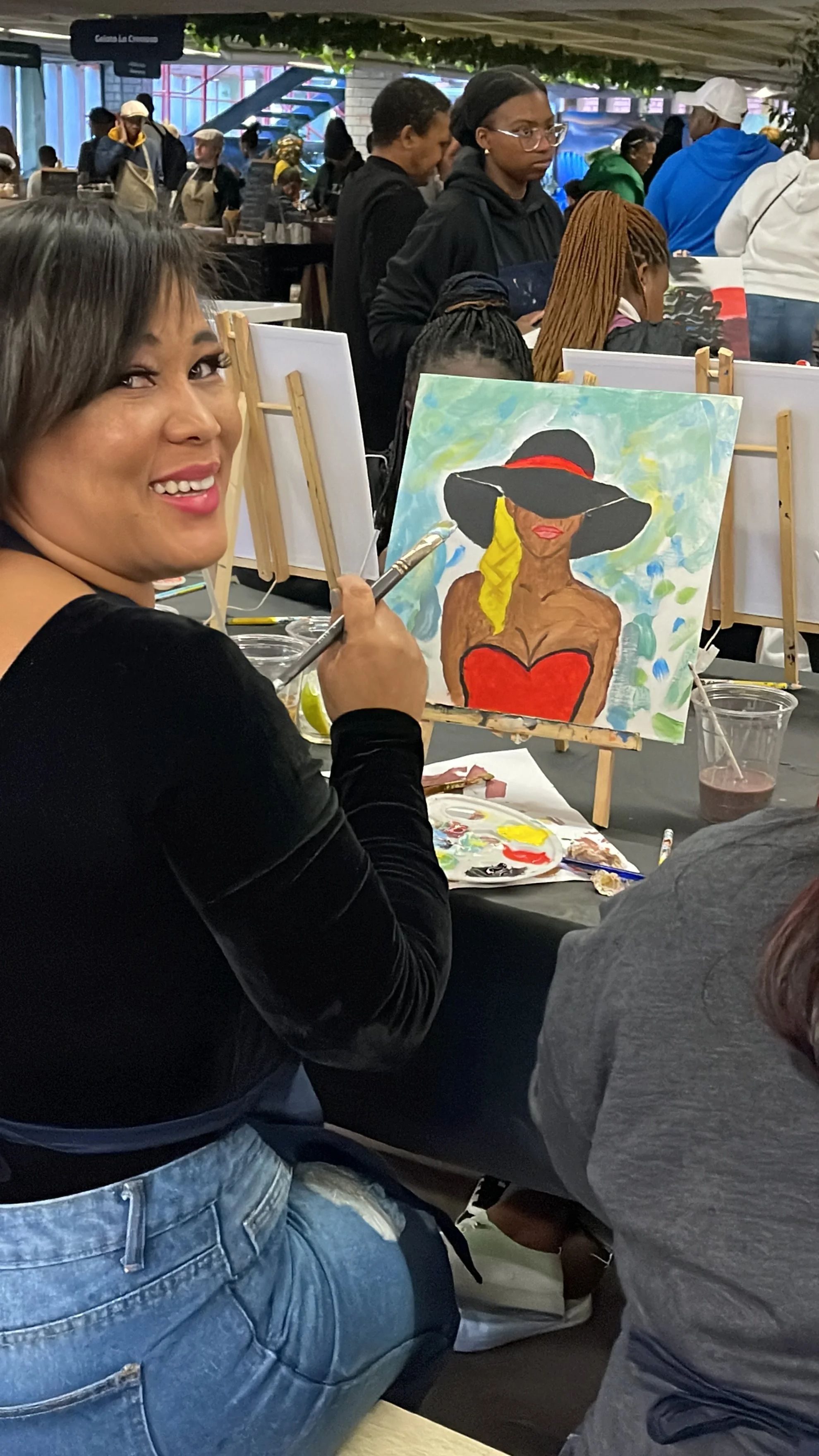 A woman with short dark hair and a black top smiling while painting a colorful portrait of a woman wearing a large hat, red dress, and yellow scarf, seated at an art event with other people and paintings in the background.