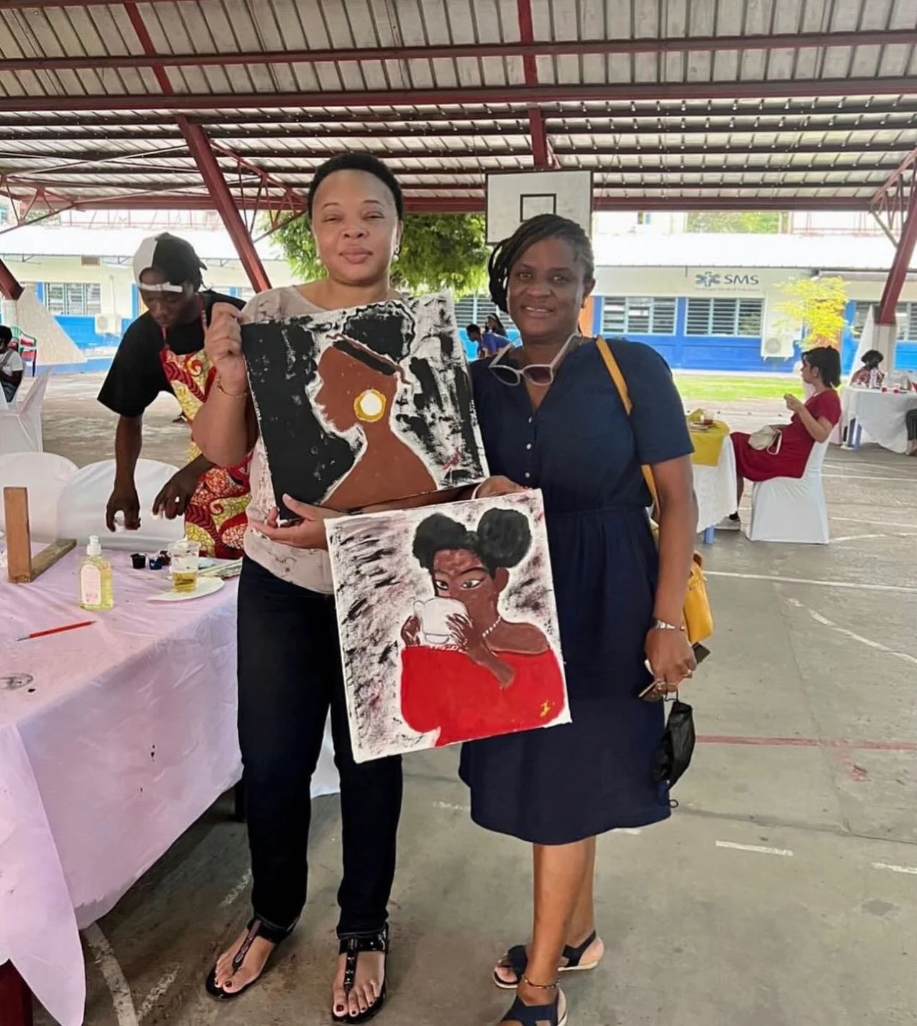 Two women standing in an indoor sports arena, holding paintings featuring stylish women. One woman wears a beige top and black pants, and the other wears a navy blue dress. There are people sitting and standing in the background, and a table with art