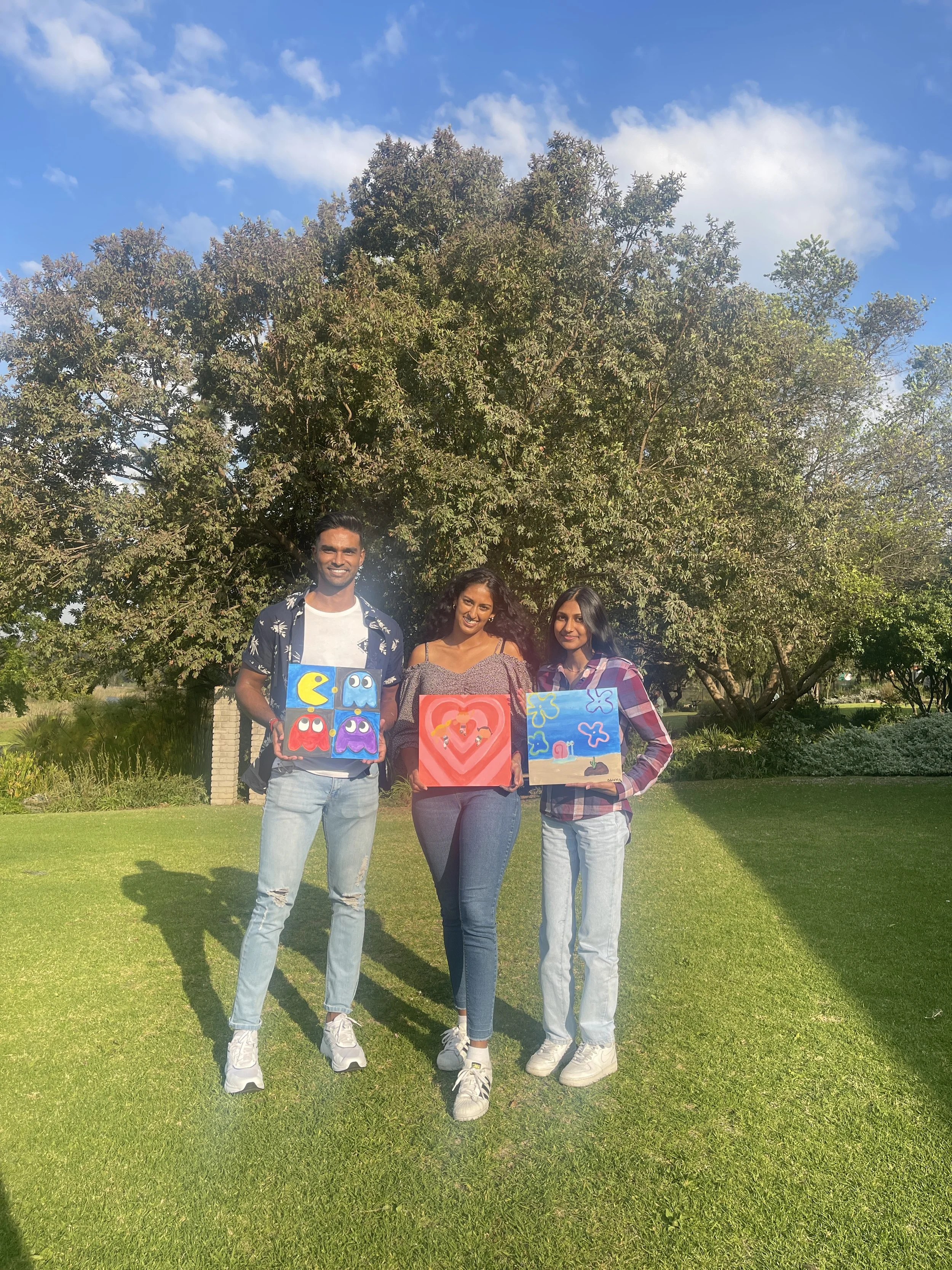 Three people standing on grass holding colorful paintings with a large tree and blue sky in the background.