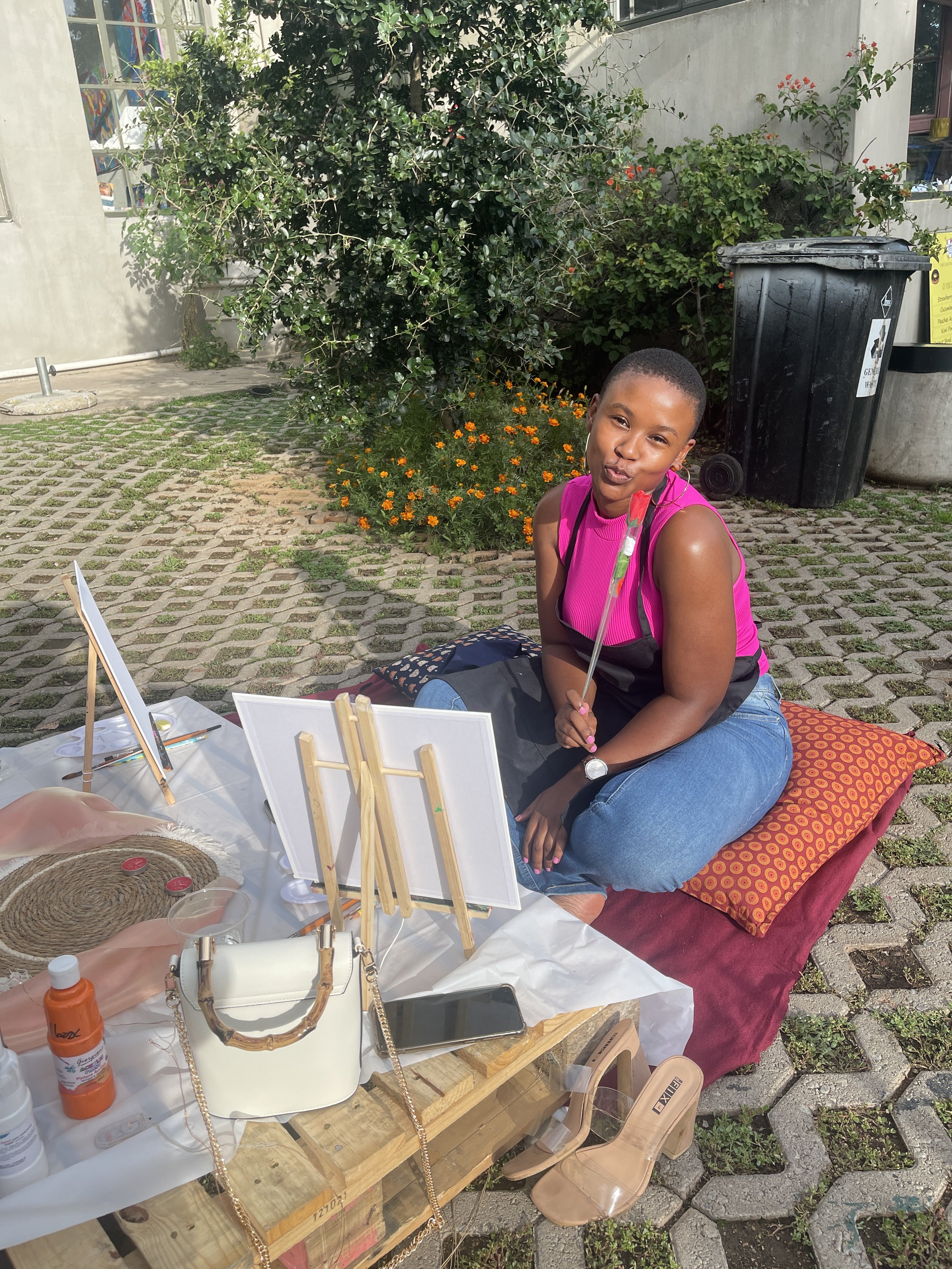 A young woman with short hair, wearing a pink sleeveless top and blue jeans, sitting cross-legged on a red cushion outside on a brick and concrete patio. She is holding a paintbrush and appears to be painting. There are art supplies on a white table 