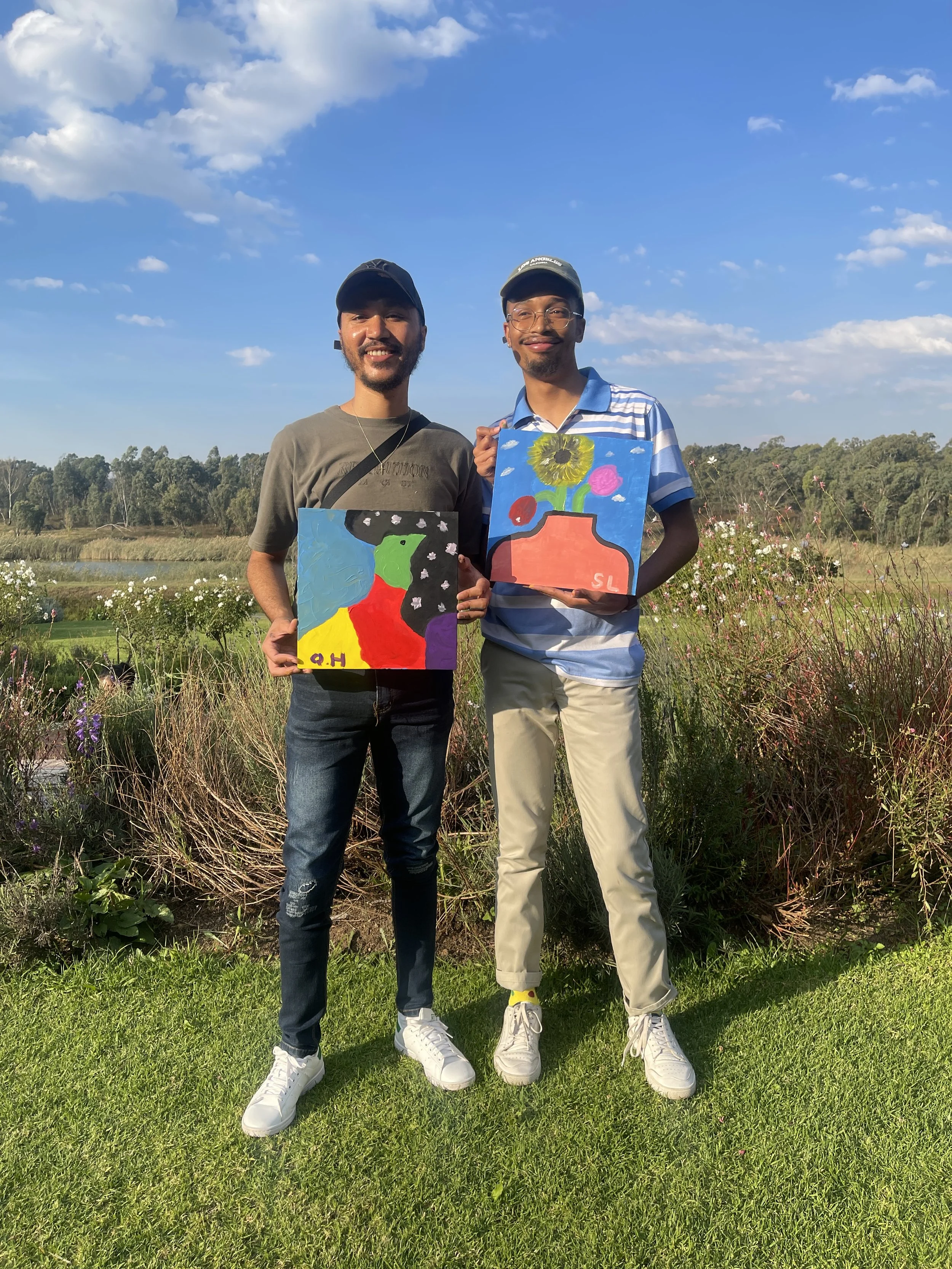 Two young men standing outdoors on a sunny day, holding colorful paintings, with a grassy field, wildflowers, and trees in the background.