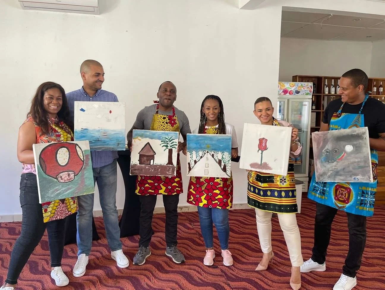 Group of six people standing indoors, holding paintings they created, and smiling for a photo. The paintings depict various colorful scenes and objects.