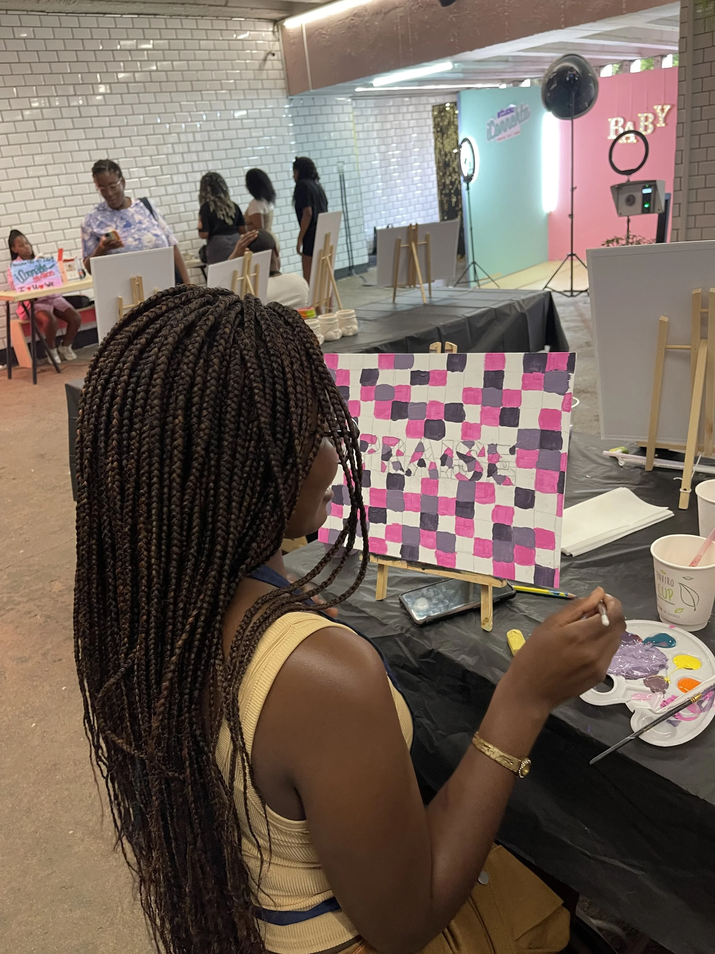 A woman with long braided hair in a yellow sleeveless top is painting on a canvas with pink, purple, and white checkered pattern at an art event. There are other people in the background, some standing in line, and an art setup area with a photo boot