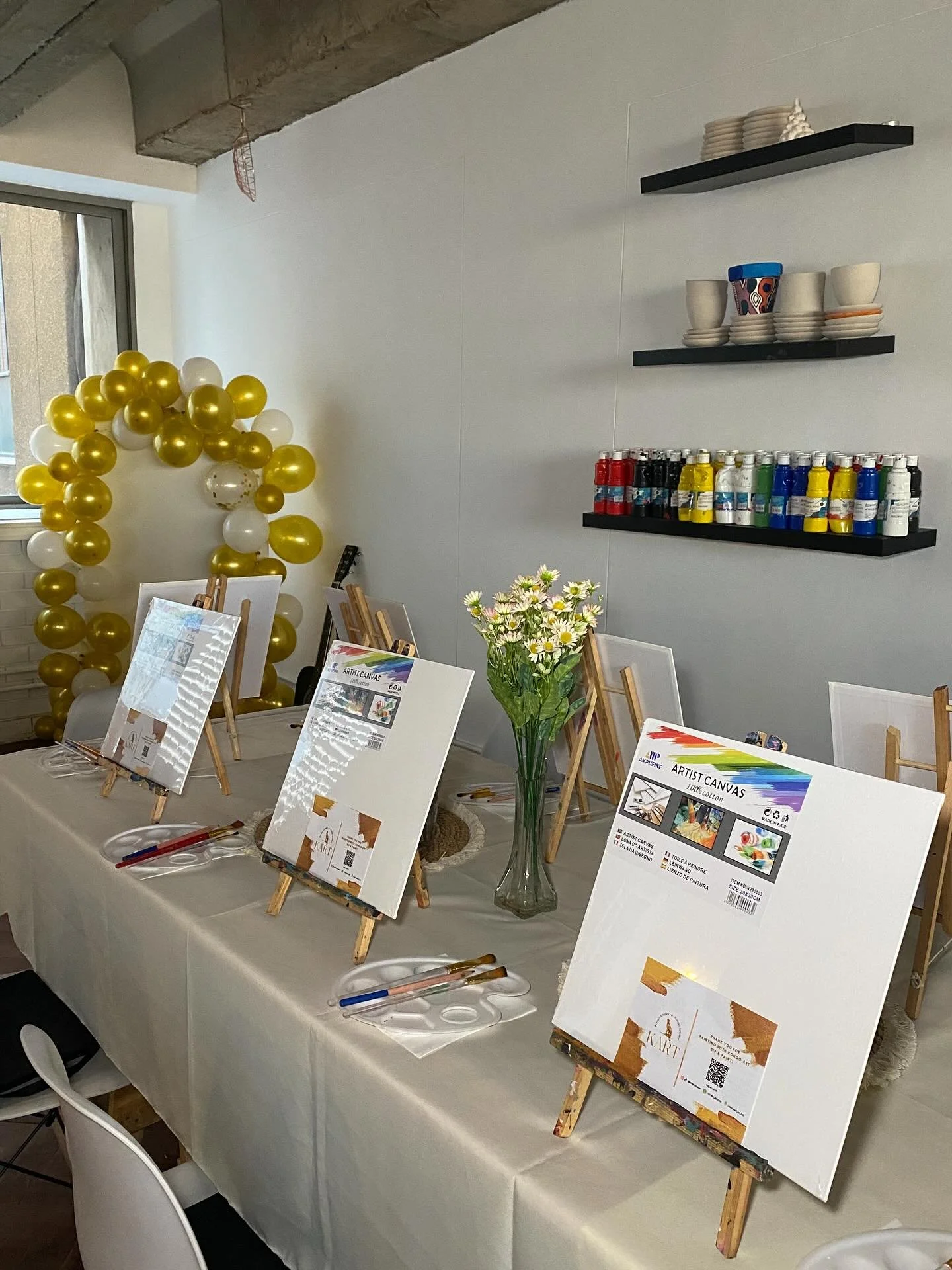 Table with art supplies, small canvases on easels, a vase of daisies, and informational posters about art canvases in a decorated indoor space.