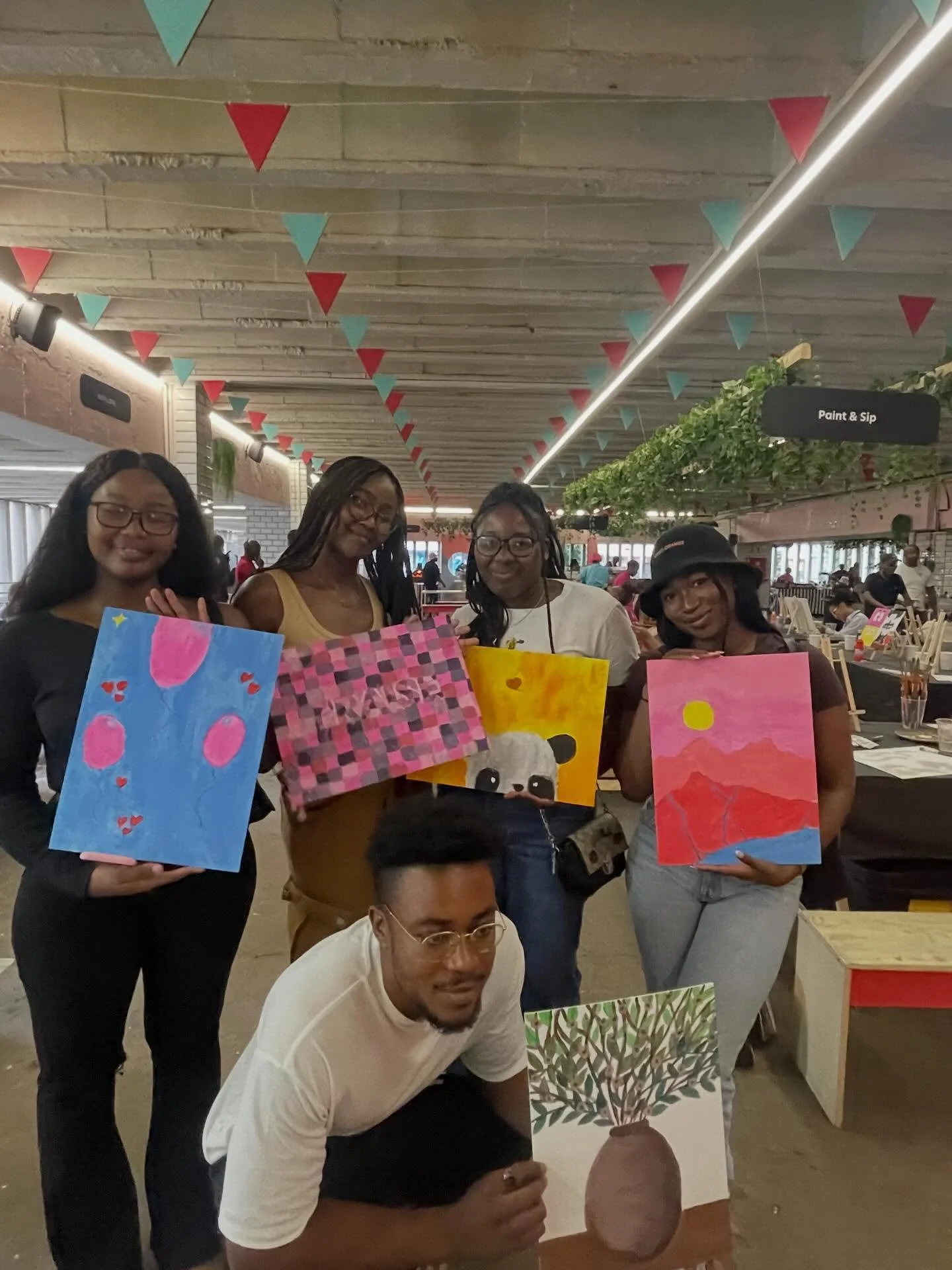 Four women and two men at an art event, holding colorful paintings of abstract and nature-themed designs, with decorations hanging from the ceiling and other people in the background.