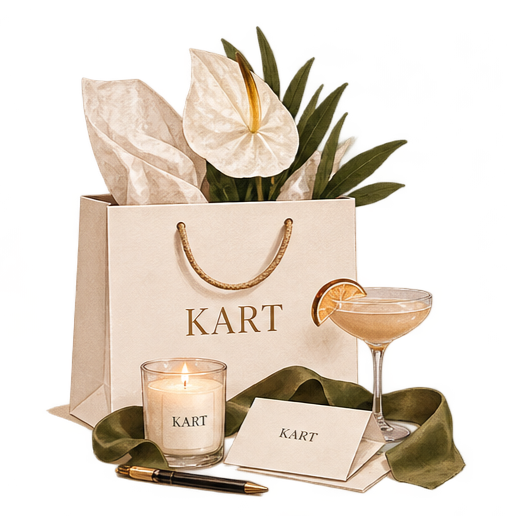 A gift bag labeled 'KART' with white tissue paper, greenery, a candle, a cocktail glass with a lemon wedge, a pen, and a card, all arranged on a green cloth.