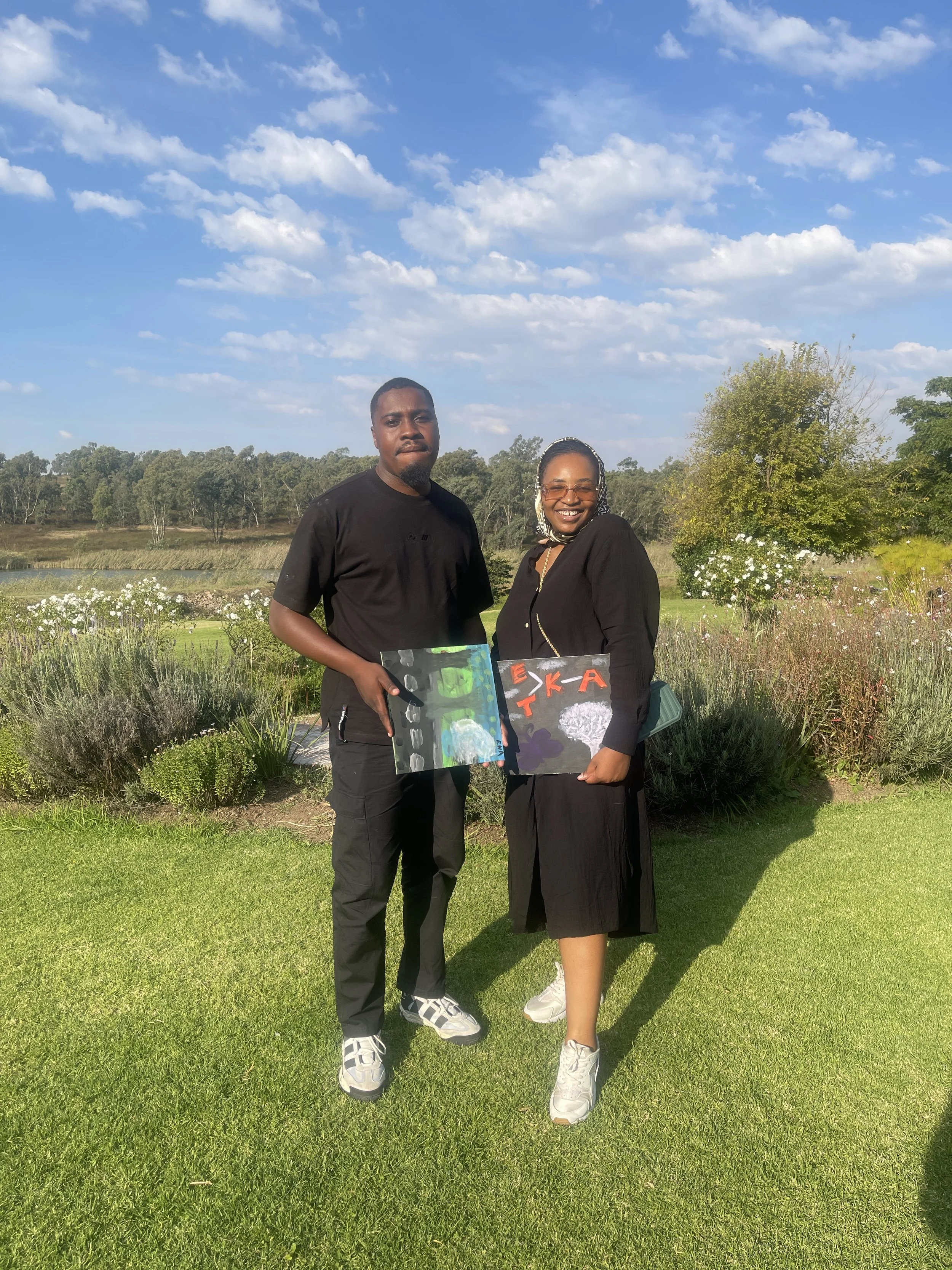 A man and woman standing outdoors on a grassy area with trees and a pond in the background, holding artwork boards, under a partly cloudy sky.
