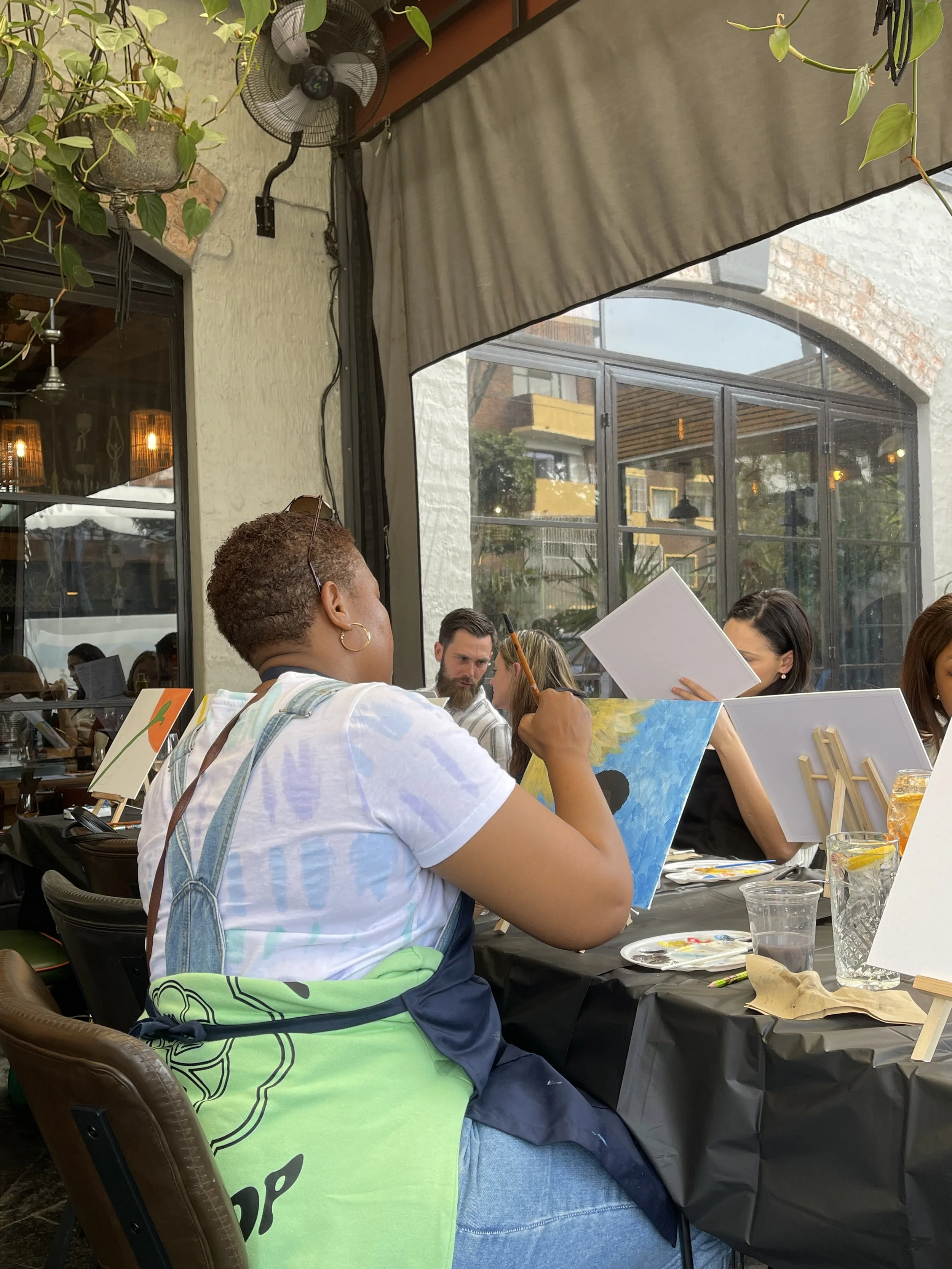 People sitting at a table with art supplies, painting canvases, and glasses of water, in an outdoor or semi-outdoor cafe with windows and plants.