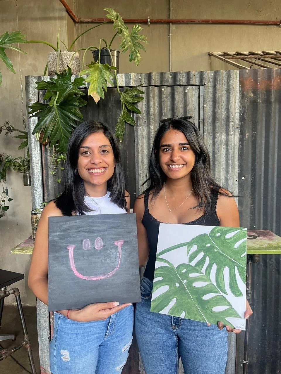 Two women standing indoors, smiling, each holding a piece of artwork: one with a gray background and a pink smiley face, the other with green tropical leaves on a white background. Potted plants and a metal wall in the background.