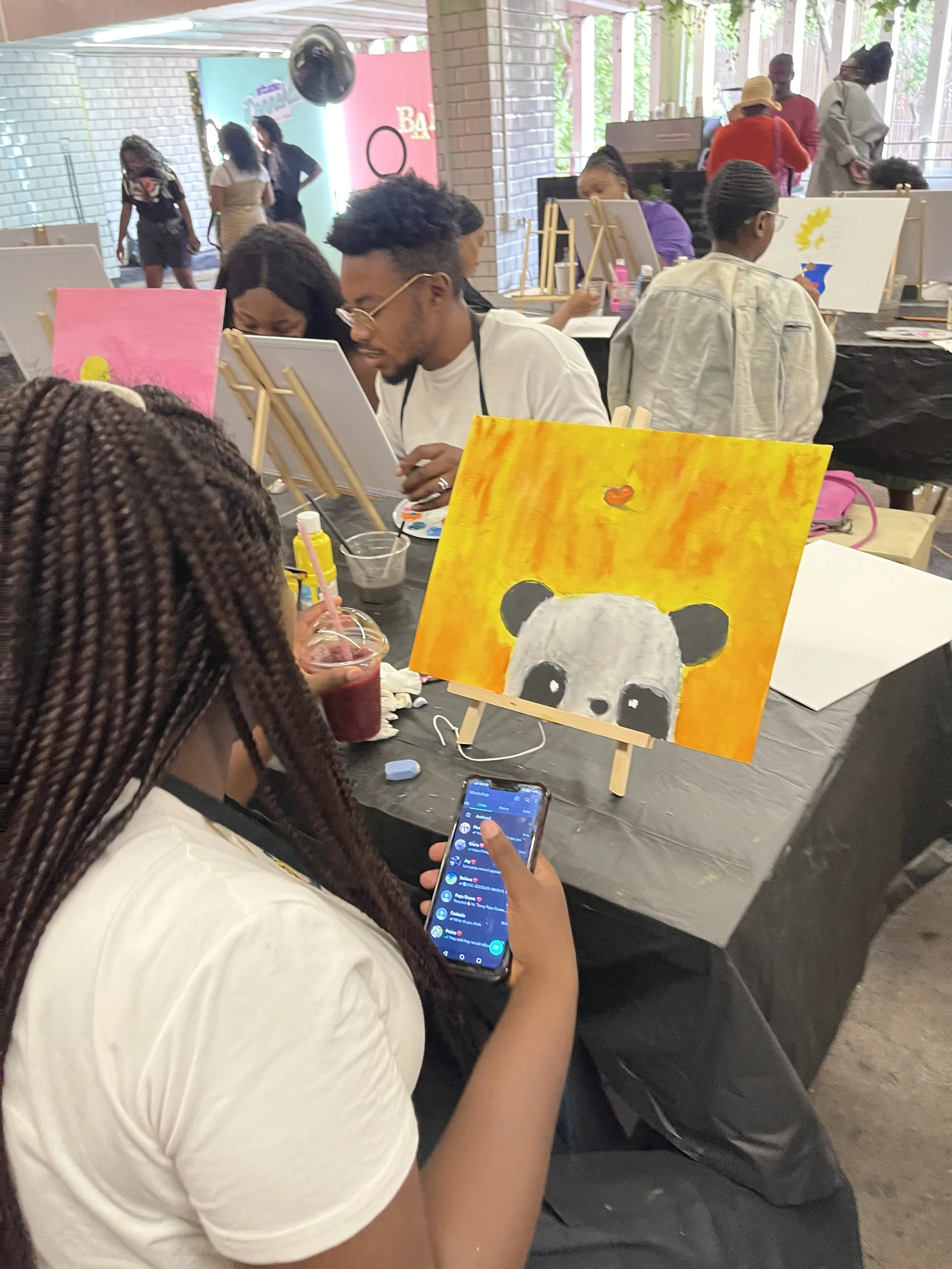People painting animal portraits at an art workshop, with paintings of pandas on easels, in a bright indoor space.