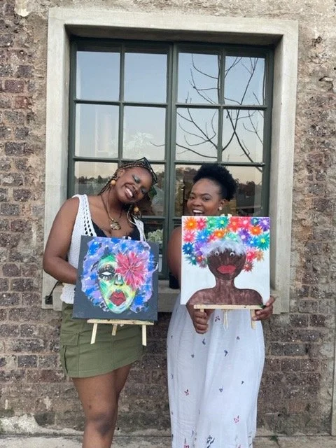 Two women holding colorful portrait paintings in front of a brick building with a window, smiling.