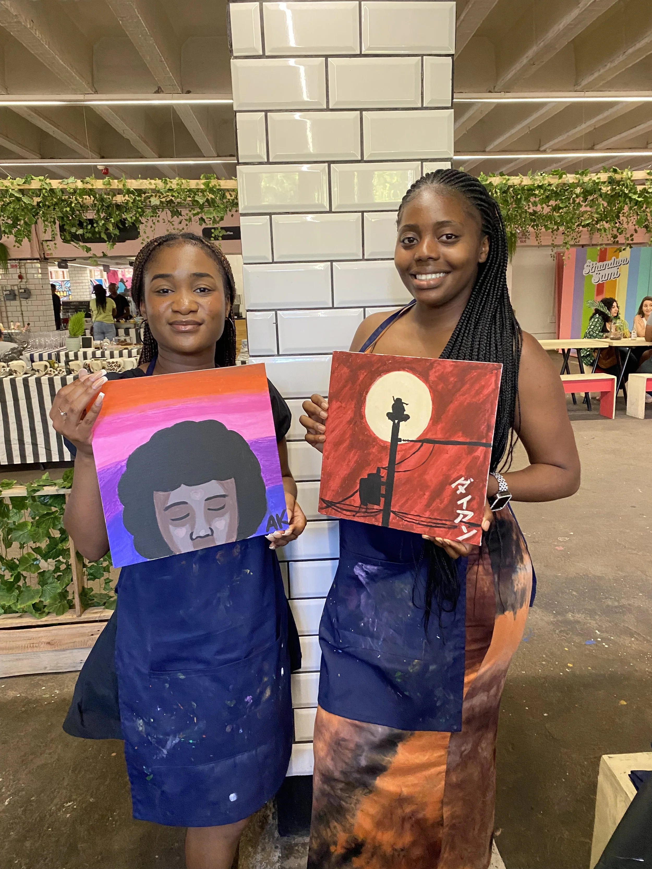 Two women standing in an indoor space, each holding a colorful painting. The woman on the left has medium skin tone, braided hair, and is wearing a dark blue apron with paint stains. The woman on the right has dark skin tone, braided hair, and is wea