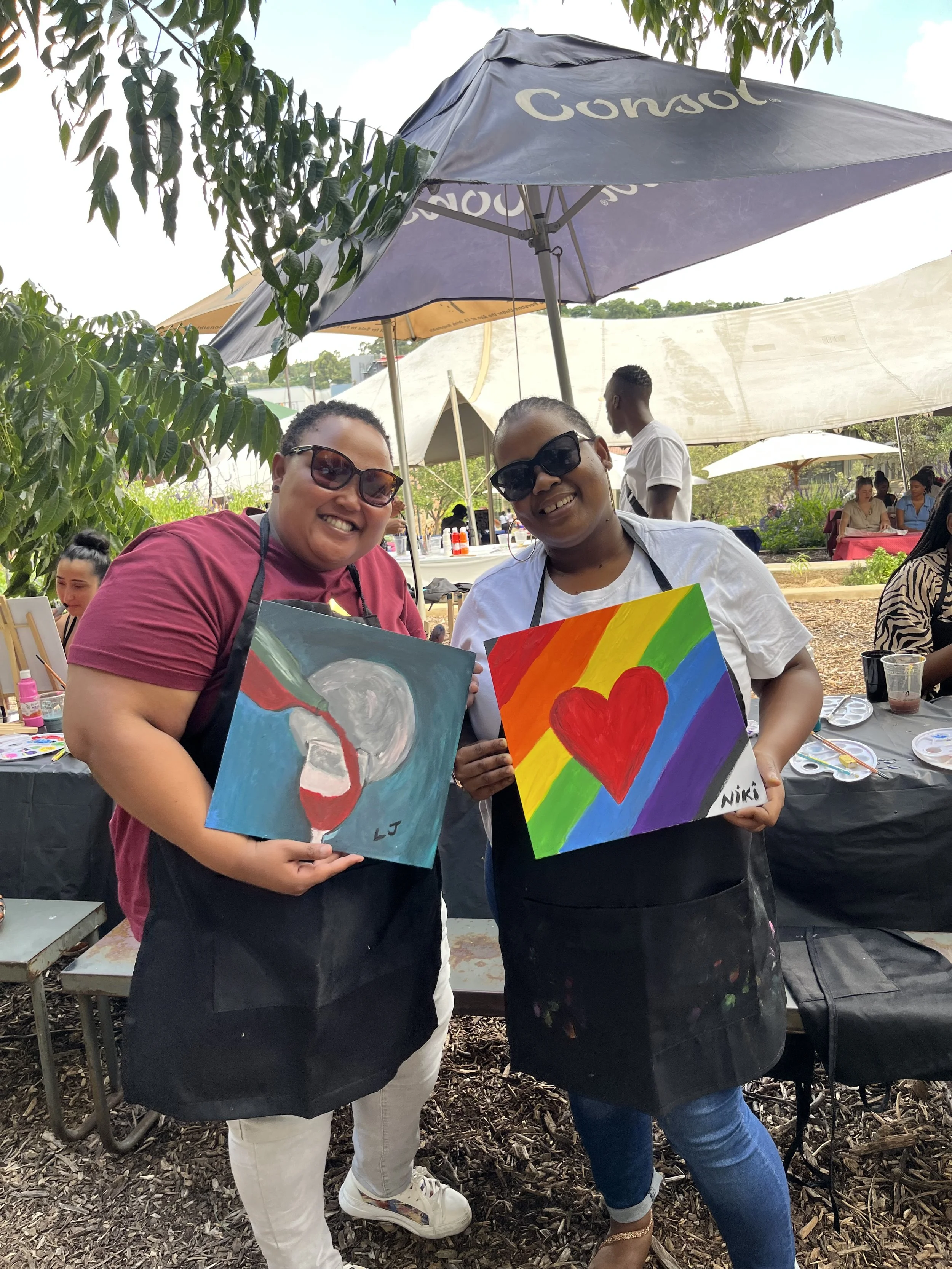 Two women smiling and holding rainbow-colored paintings at an outdoor art event under umbrellas, with other people and trees in the background.