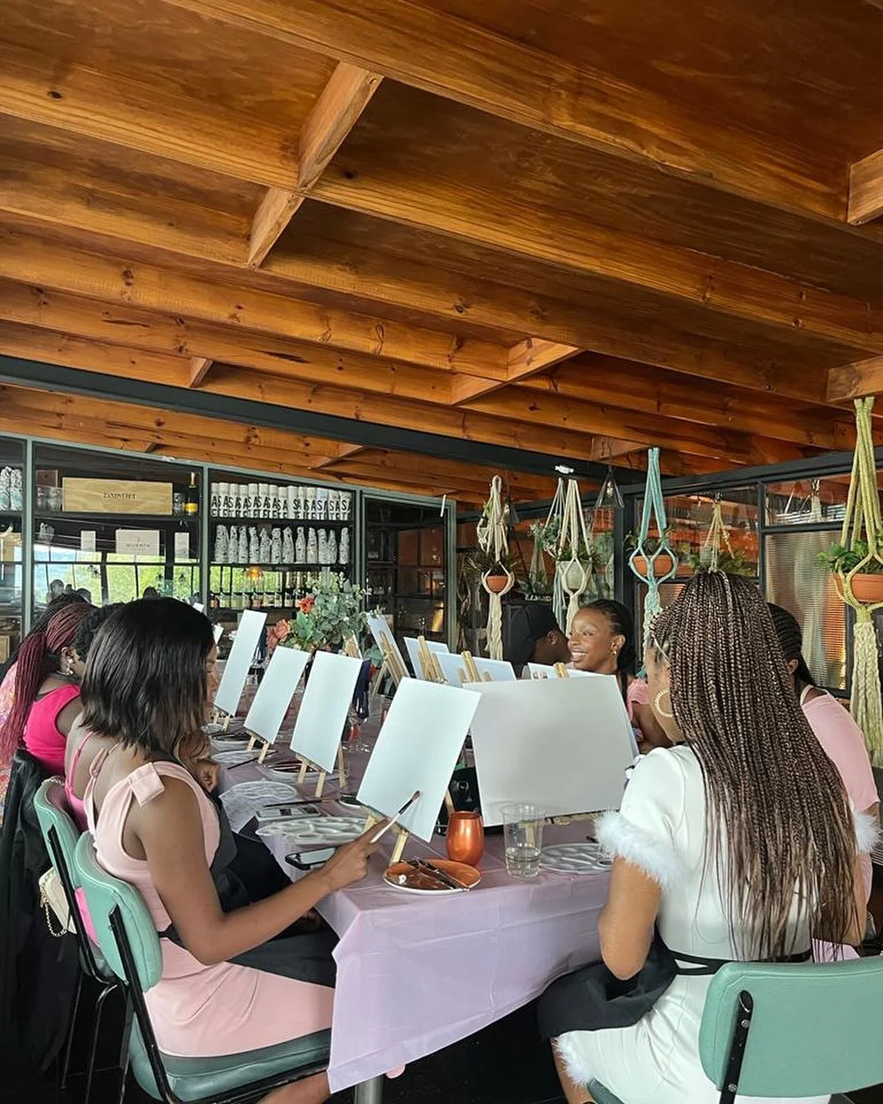 Group of women sitting at a long table, engaged in a painting activity with canvases and brushes, in a room with a wooden ceiling, hanging plants, and a windowed wall in the background.