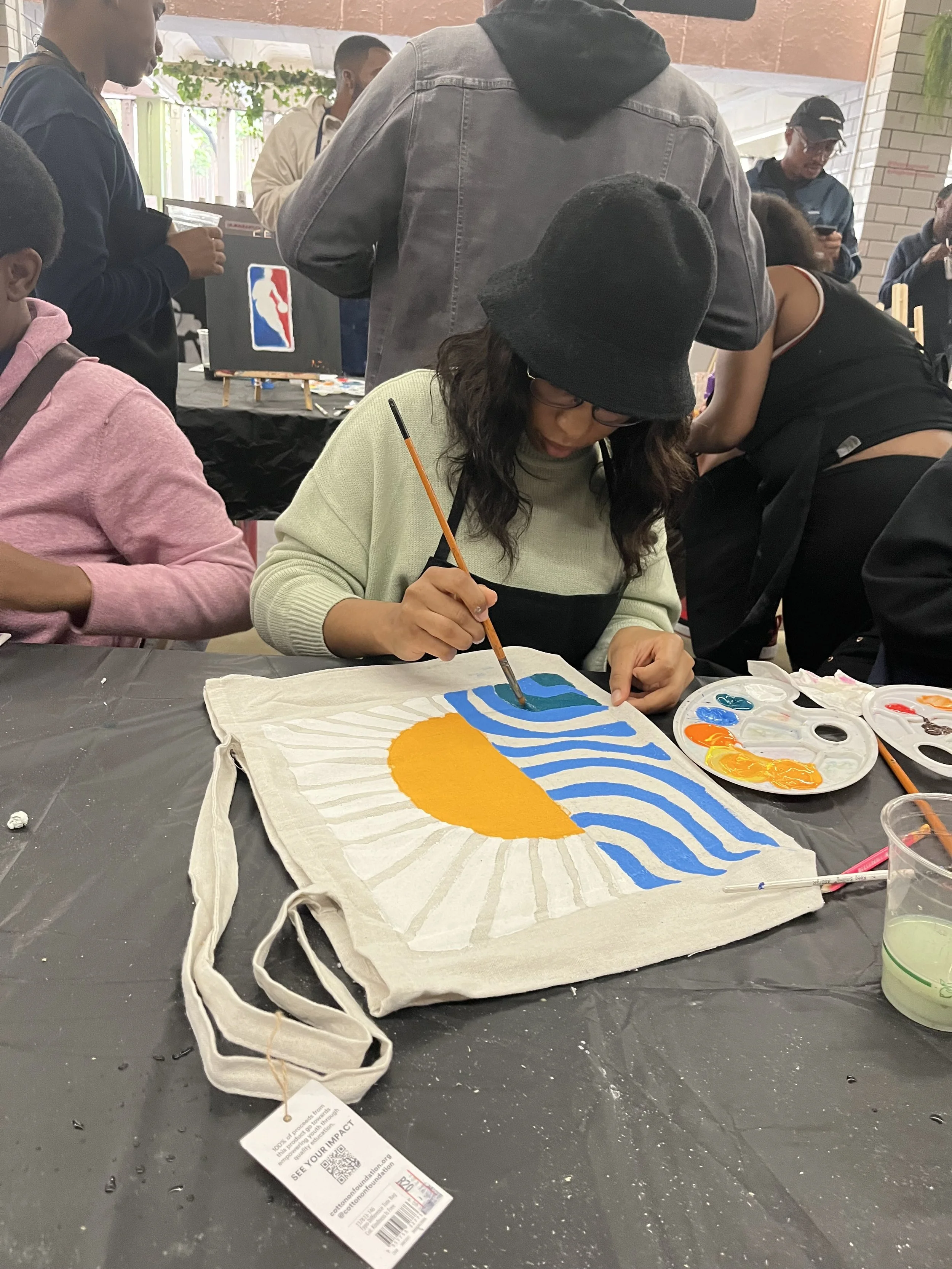 A woman wearing glasses and a black beanie is painting a design on a fabric tote bag with a sun and wave pattern using orange, yellow, and blue paints. The bag is on a black table with paint palettes and brushes. Several people are gathered around, s