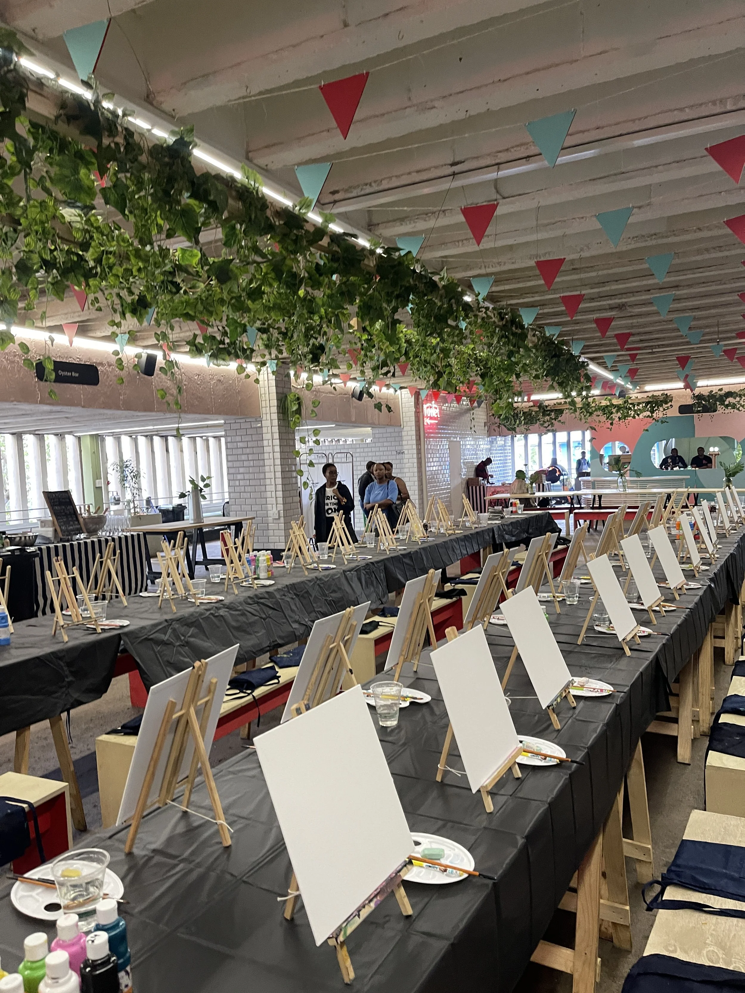 Indoor space decorated for an art event, featuring tables with easels, blank canvases, and art supplies, hanging red and blue bunting, green plants, and several people preparing or working at the tables.