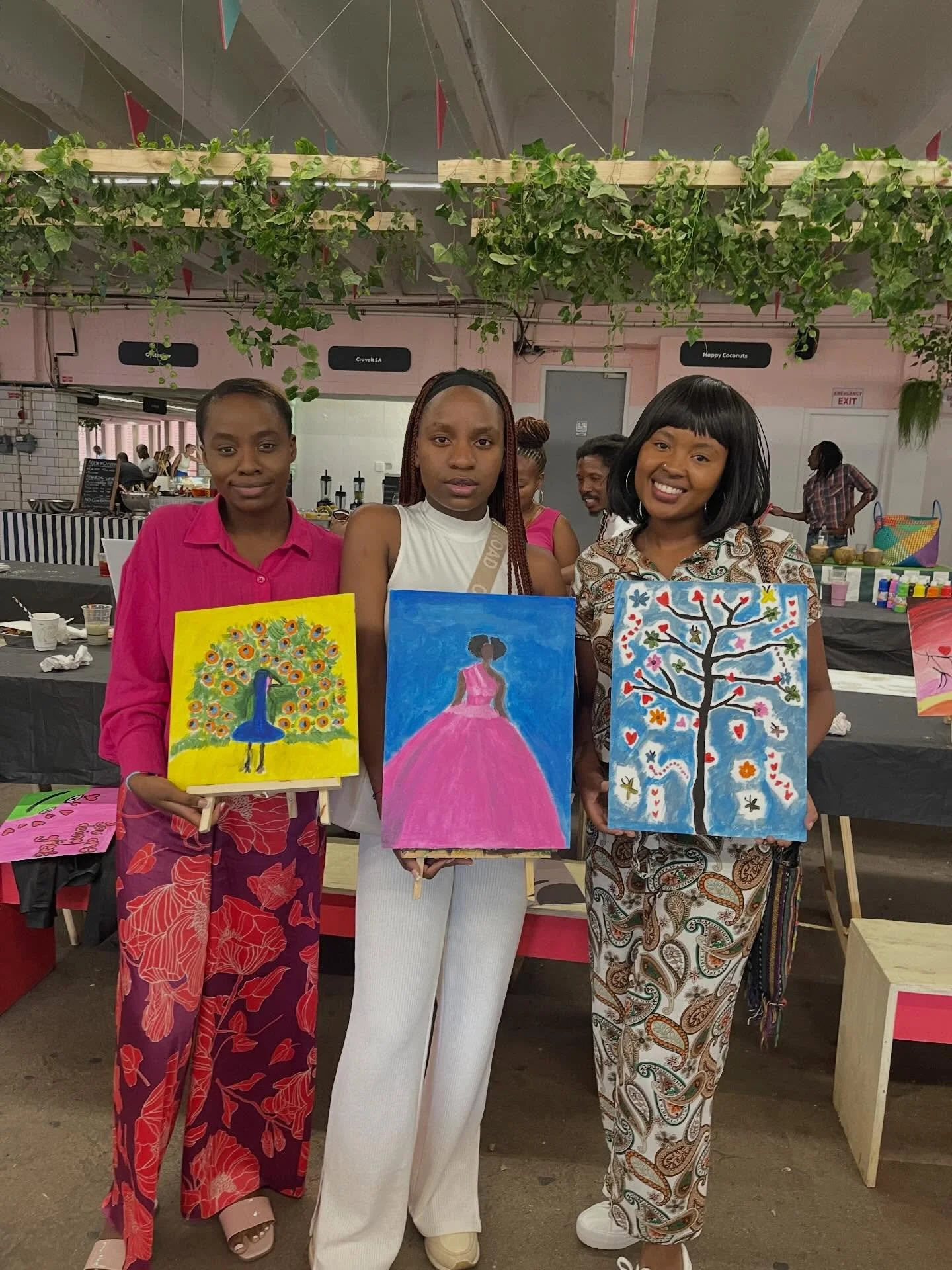 Three women standing together at an art event, each holding a colorful painting of trees.