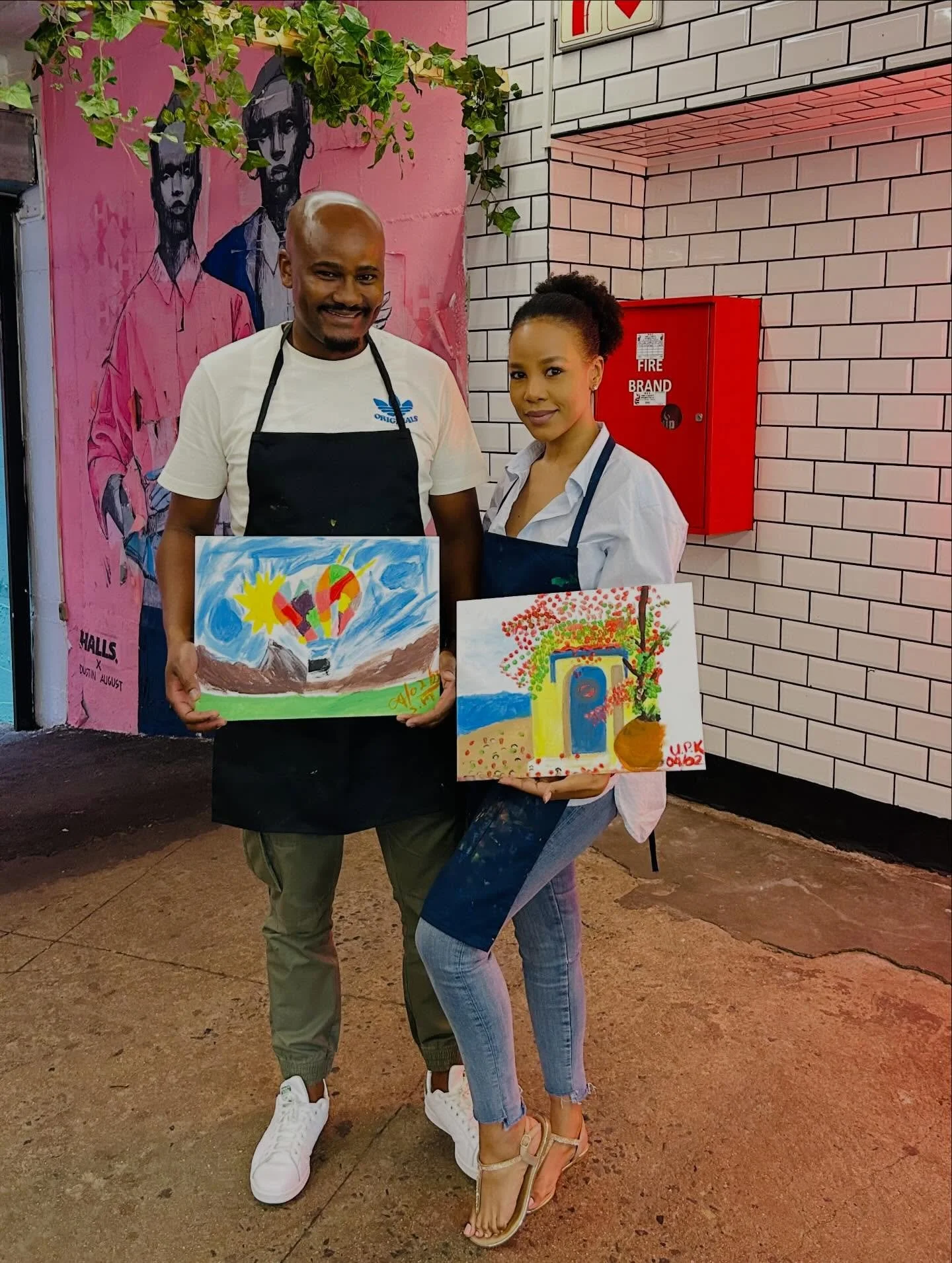 A man and woman holding colorful paintings, standing in front of a pink mural with outlined portraits of two women and a white brick wall background, both wearing aprons and smiling.