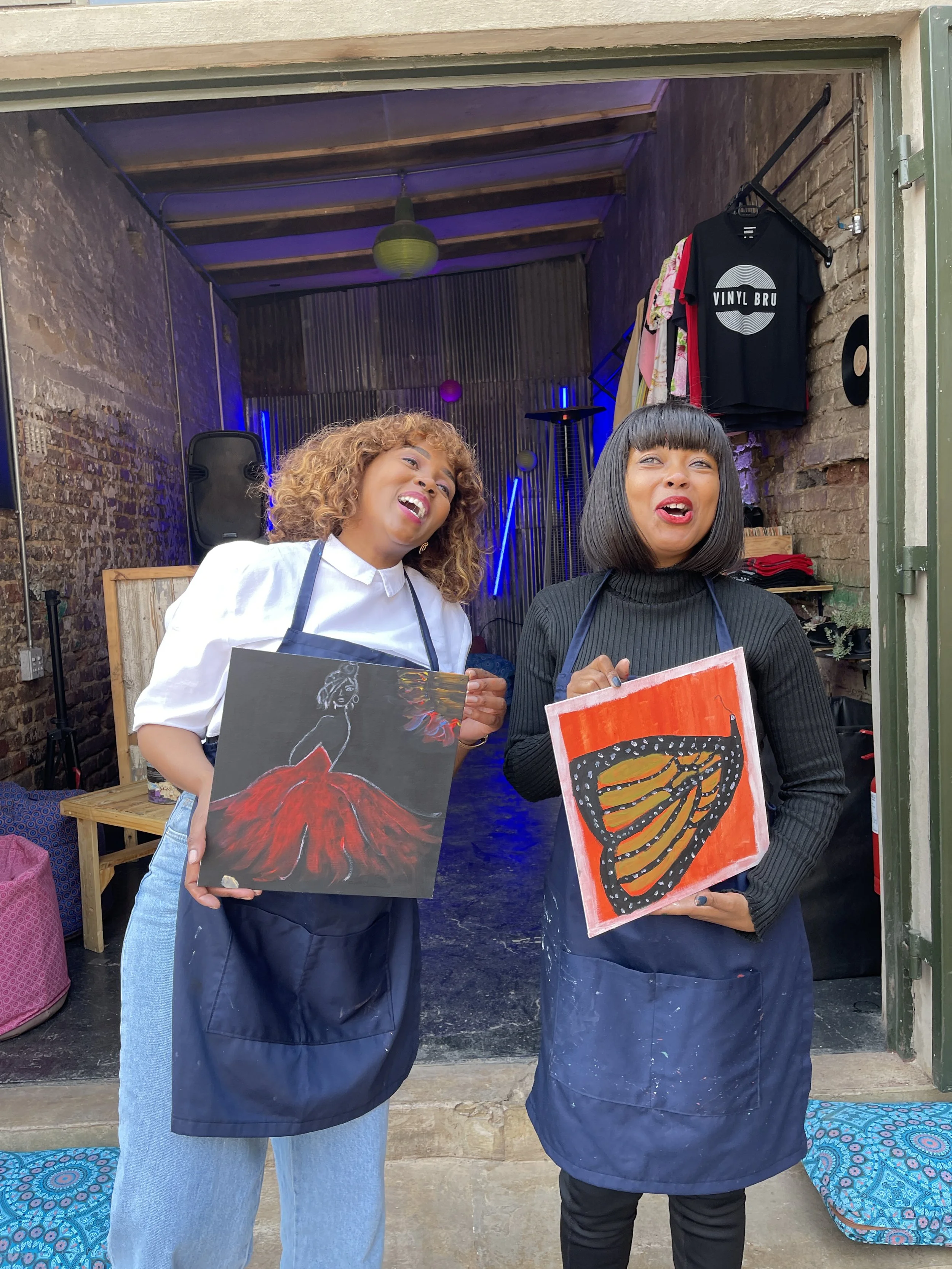Two women standing outside, each holding a painting of butterflies; one butterfly is orange and black, the other is red and black.