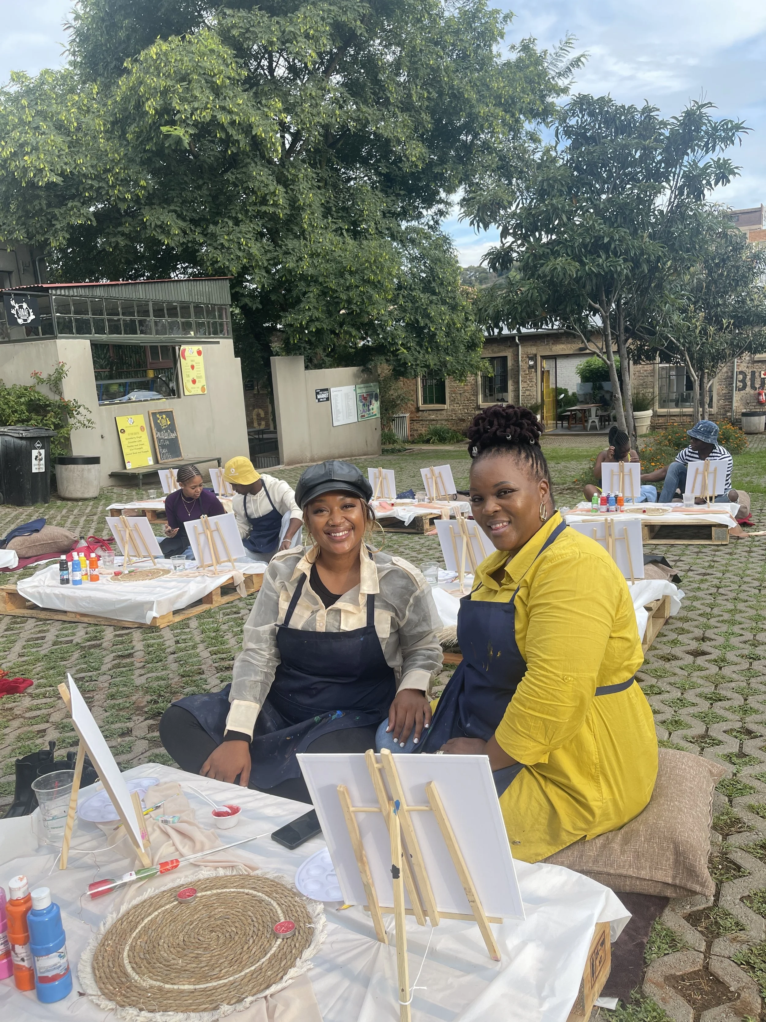 Two women sitting at an outdoor painting event, surrounded by painting supplies, with other participants and trees in the background.
