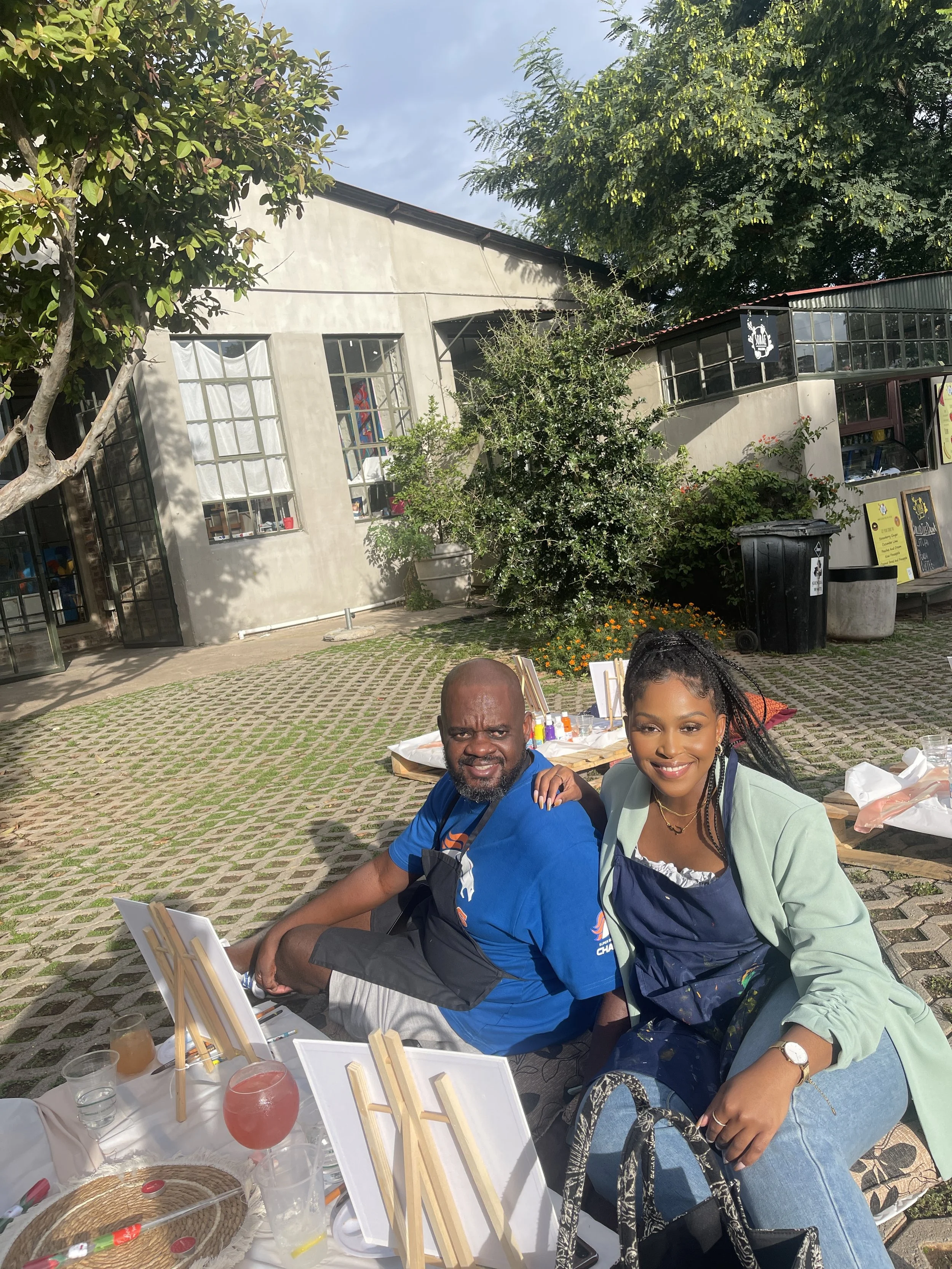 Two people sitting at an outdoor event table, smiling, with art supplies and drinks in front of them, surrounded by trees and a building in the background.