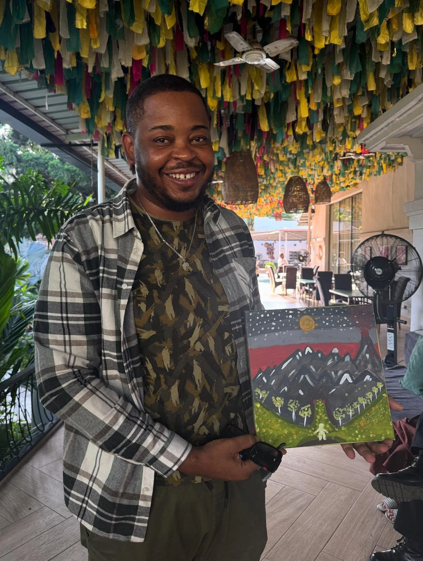 A smiling man holding a colorful painting, standing inside a decorated venue with hanging fabric and lighting.