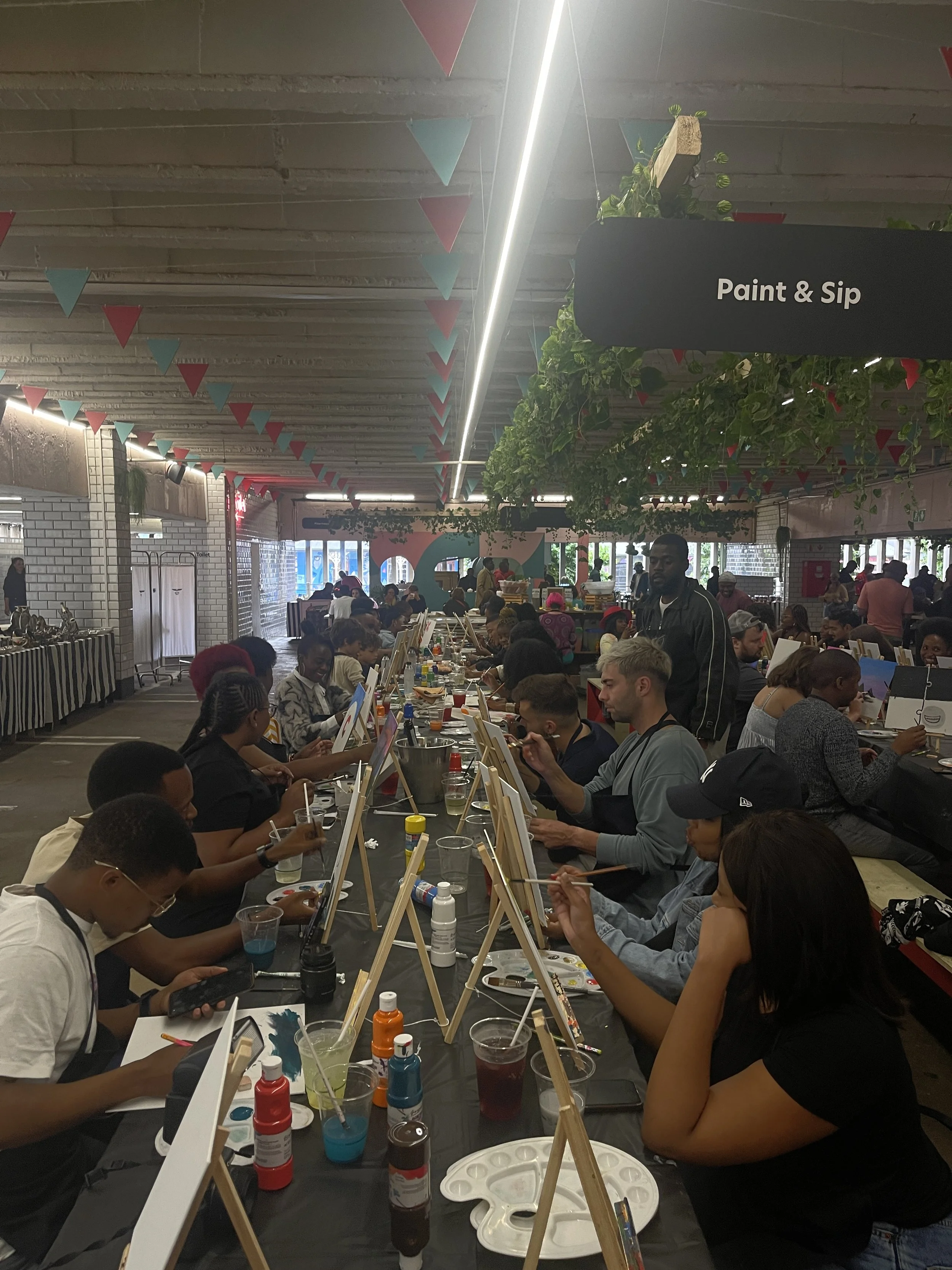 People sitting at long tables painting on canvases in an indoor art workshop with paint bottles, brushes, and palettes, decorated with colorful hanging triangular flags and greenery.